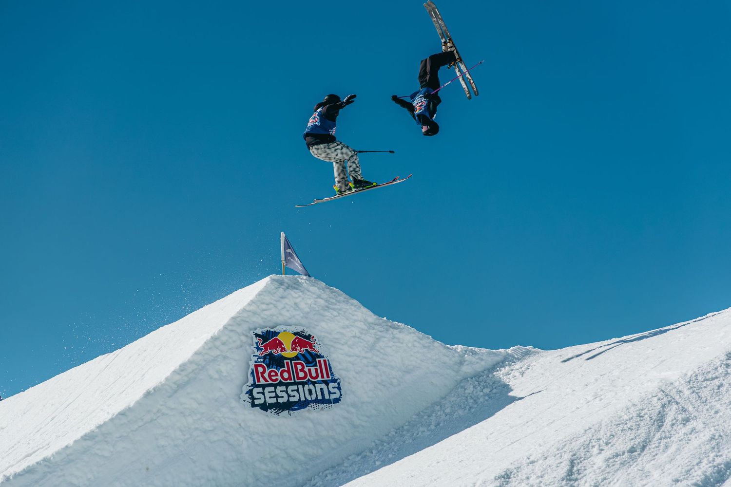 Two skiers perform aerial tricks over a snow jump at a Red Bull Sessions event under a clear blue sky.