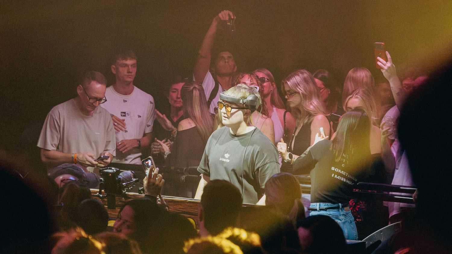 Person wearing VR headset performing on stage surrounded by cheering crowd in a dimly lit venue.