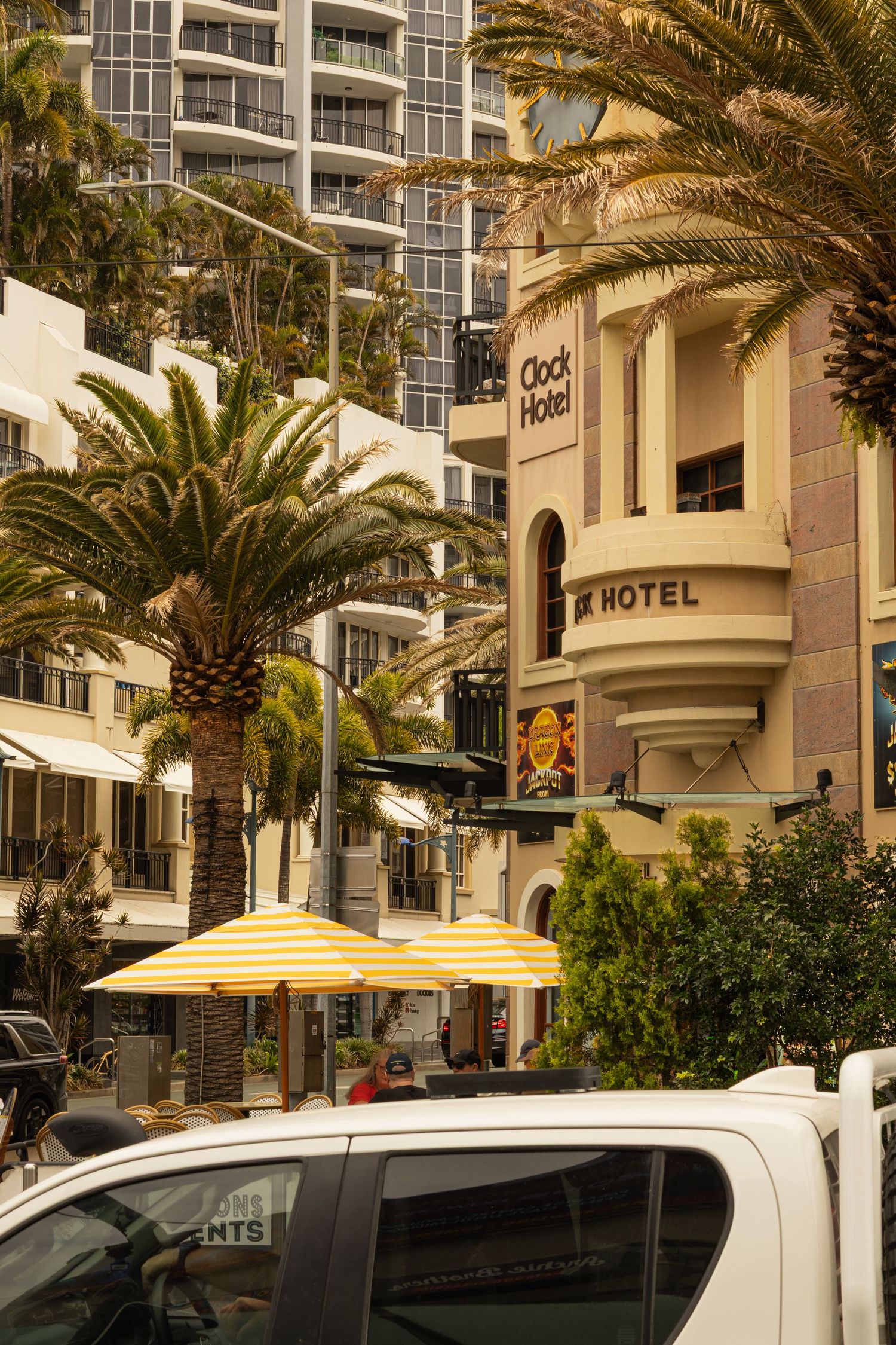 Luxury hotel exterior with palm trees and yellow umbrellas along a street lined with parked cars.