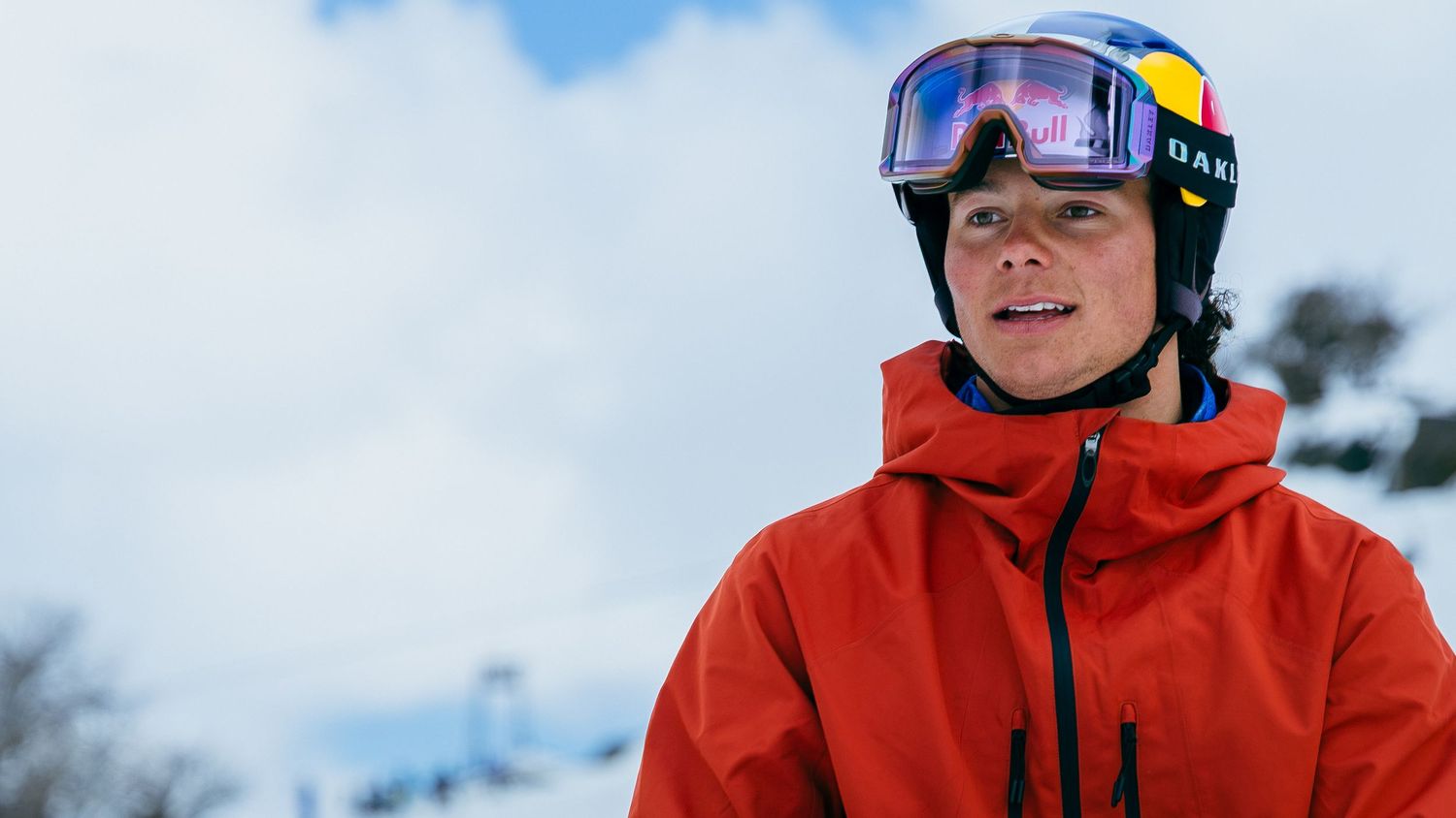 Young male skier in red jacket and Oakley goggles with Red Bull helmet poses on snowy mountain slope.