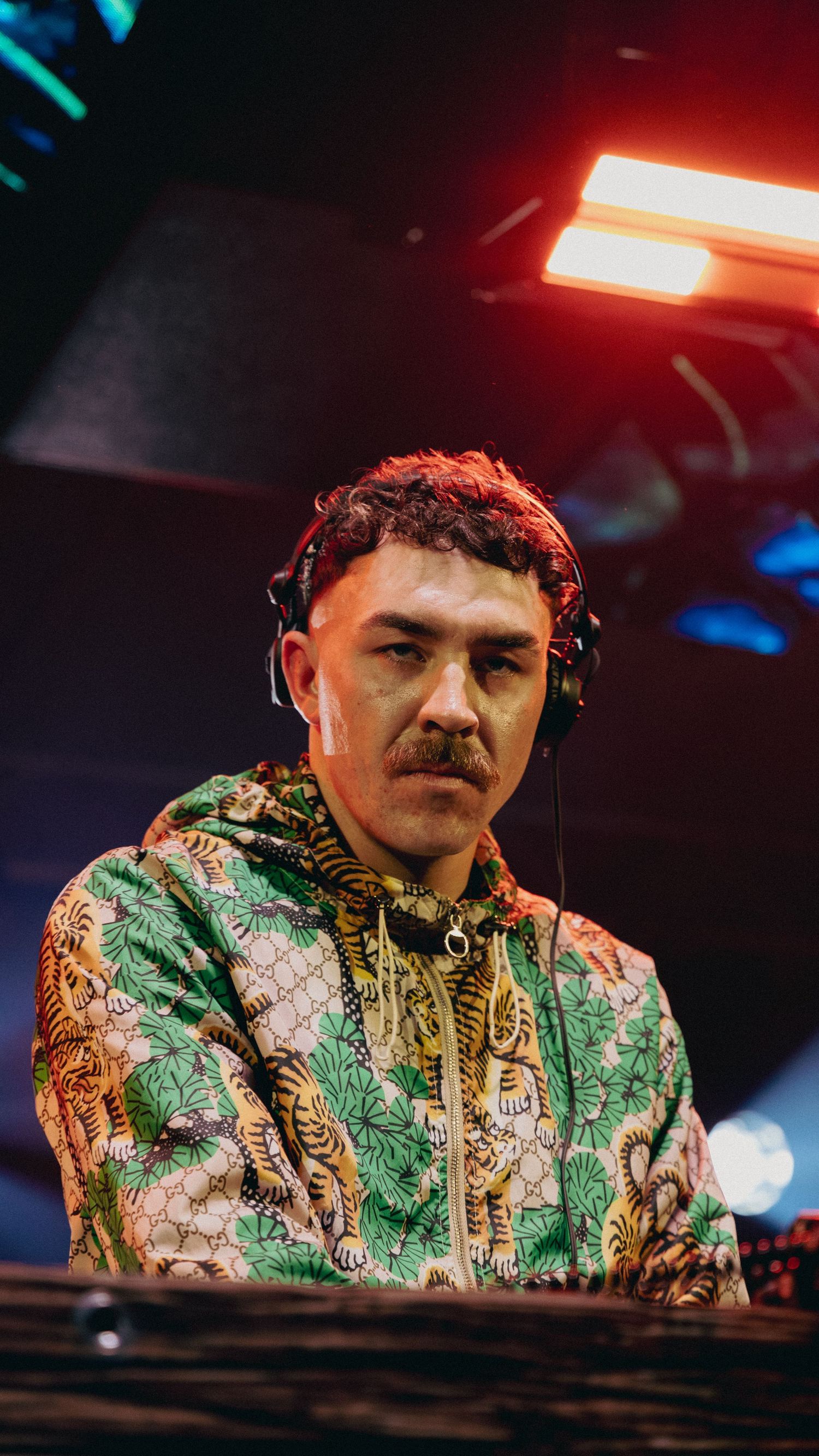 DJ wearing headphones and a colorful tiger-print Gucci hoodie performing at a nightclub under red stage lighting.