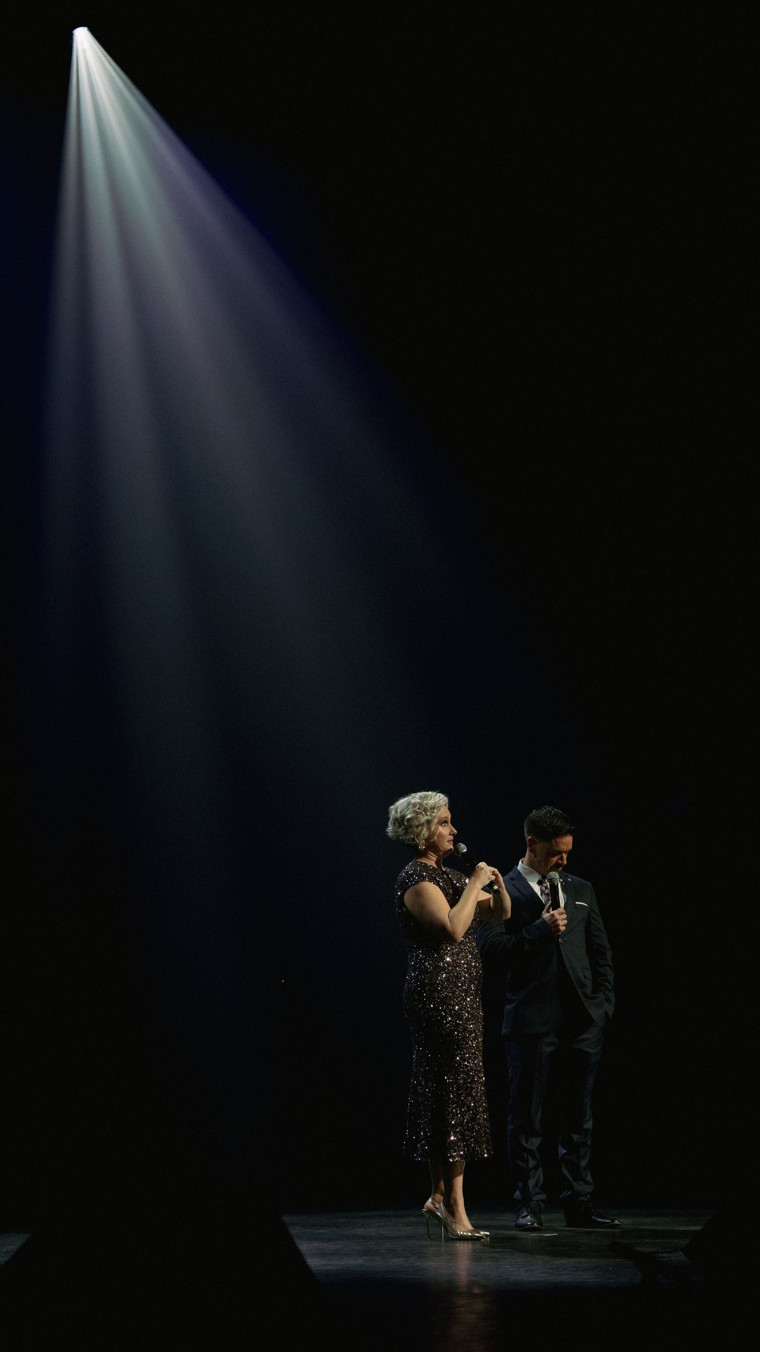 Two performers sing on a dramatic dark stage under a single spotlight during a live theater show.