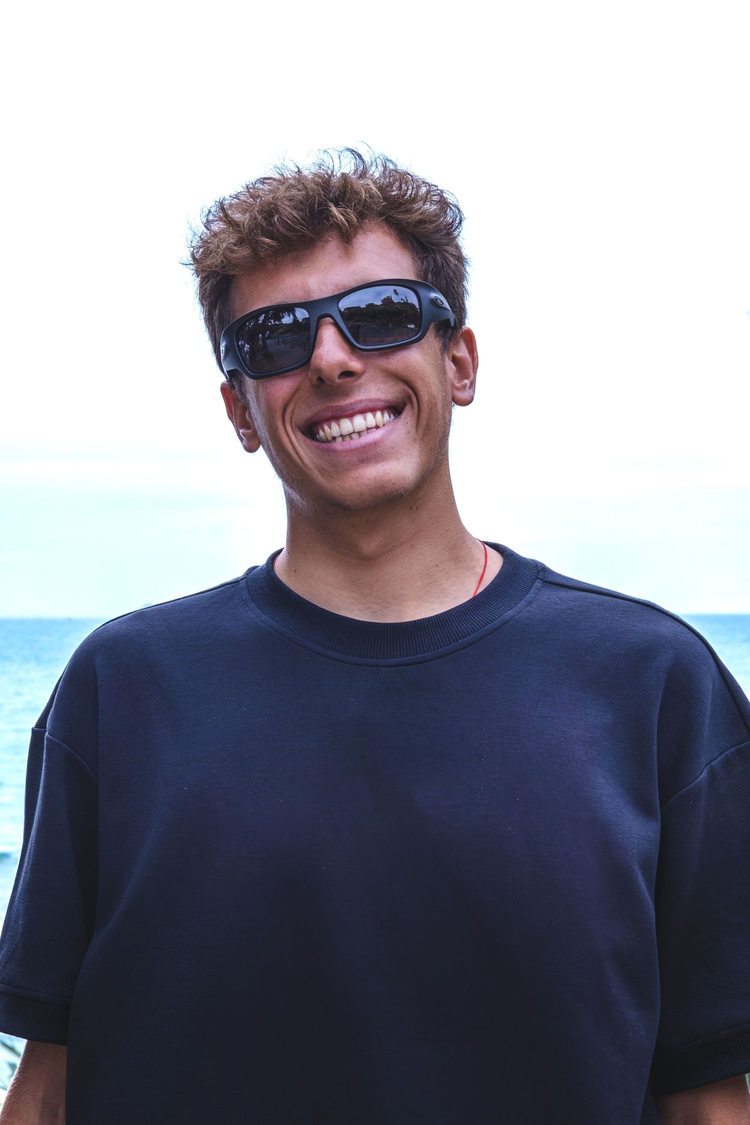 Person wearing sunglasses and a black t-shirt smiles broadly while outdoors against a bright sky background.