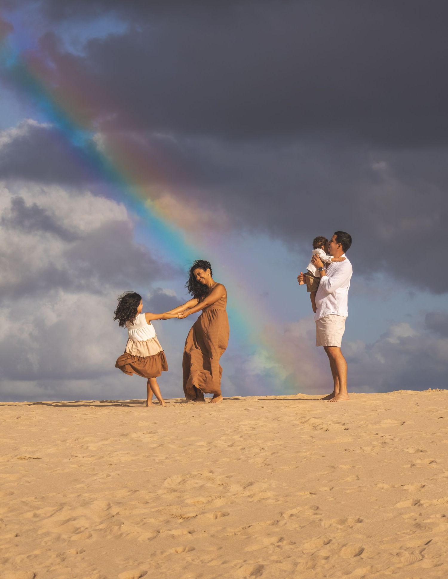 Family plays together on a sandy beach dune at sunset with a rainbow in the dramatic cloudy sky.