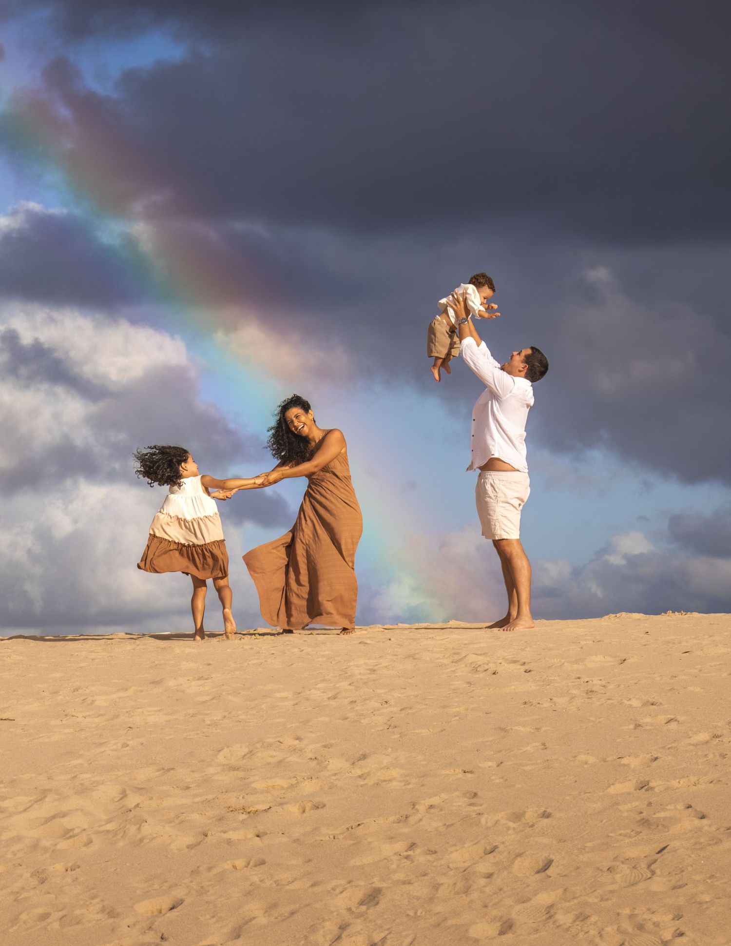 A family plays together on a sandy beach at sunset with a beautiful rainbow arcing through the dramatic cloudy sky.