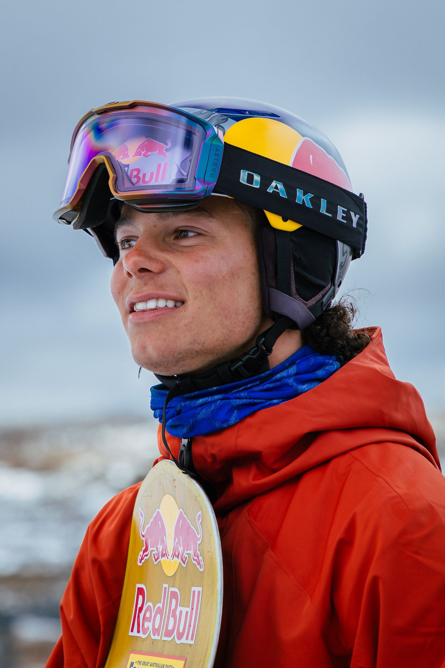 Smiling snowboarder wearing Oakley goggles, Red Bull helmet, and red jacket holds a Red Bull snowboard outdoors.