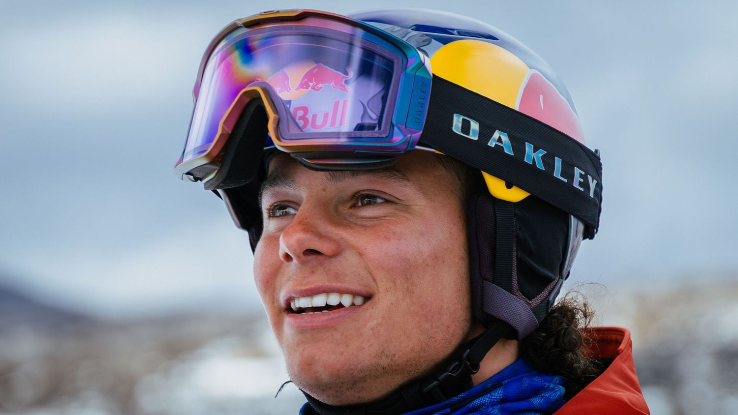 Smiling athlete wearing Oakley goggles and Red Bull helmet against a snowy mountain backdrop.