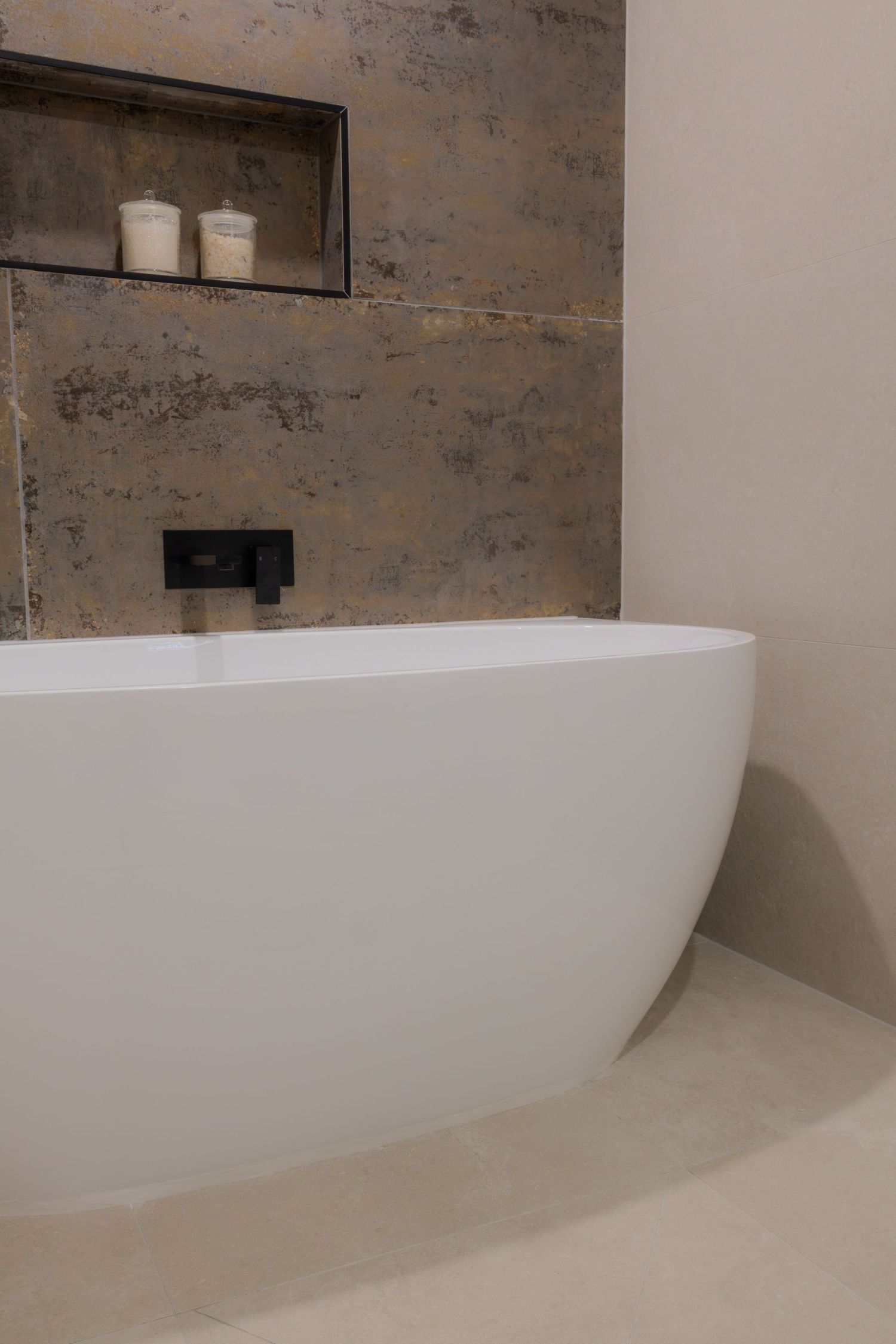 Modern freestanding white bathtub against a textured stone wall with black fixtures and a built-in niche shelf.