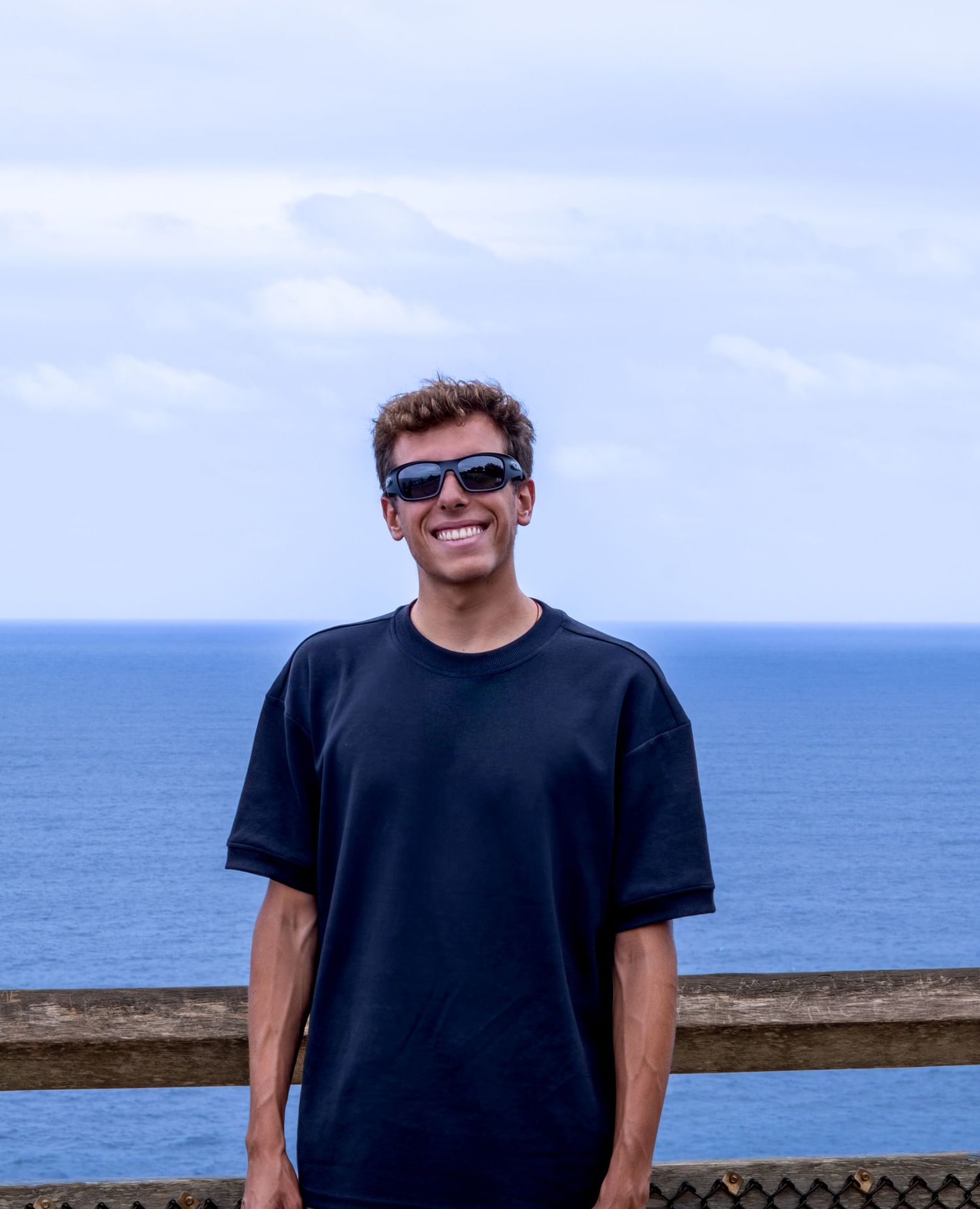 Person in a black t-shirt stands on an oceanfront walkway with blue water and sky in the background.