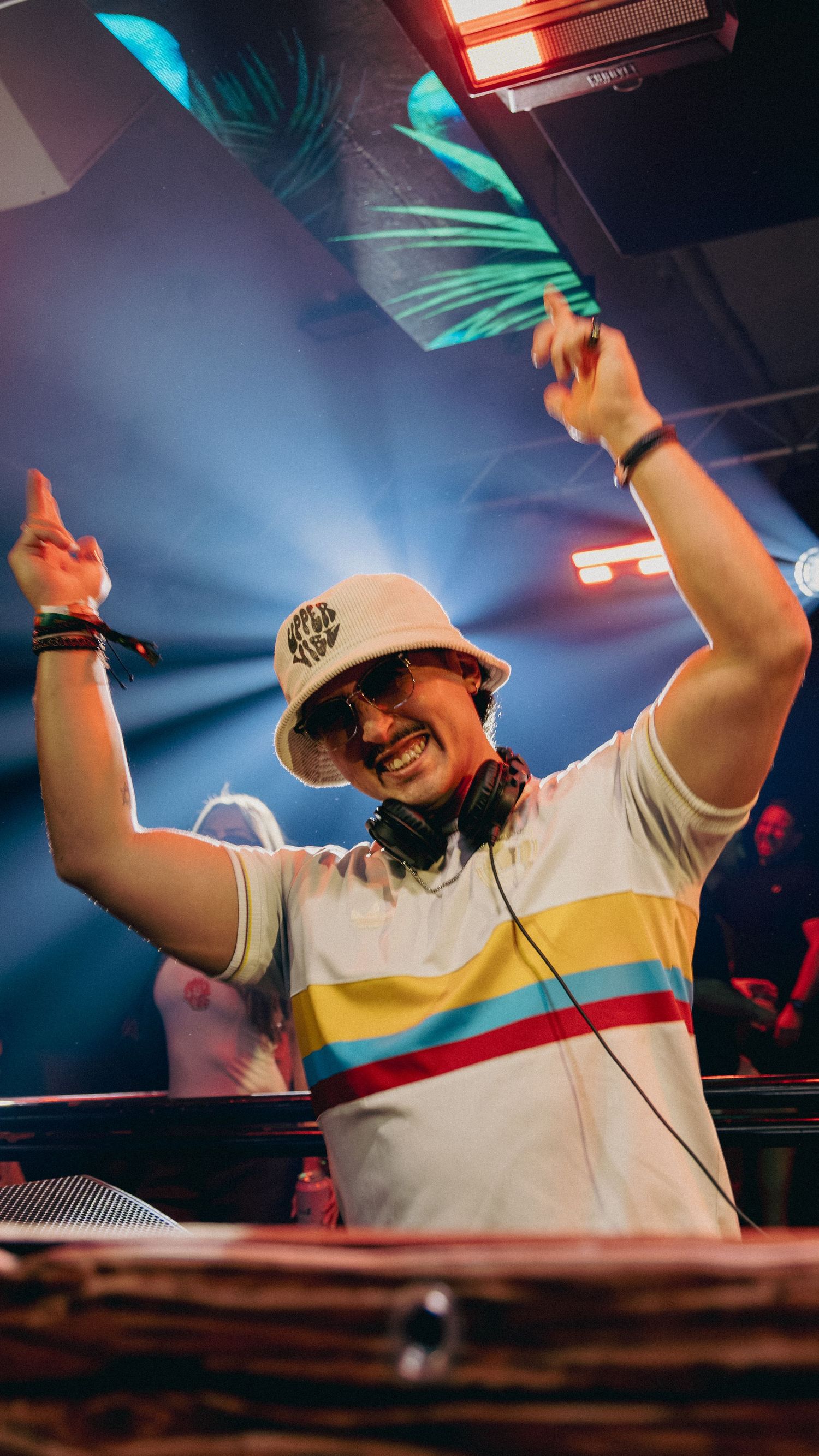 A DJ in a bucket hat and striped shirt raises hands at a vibrant nightclub with blue stage lights.