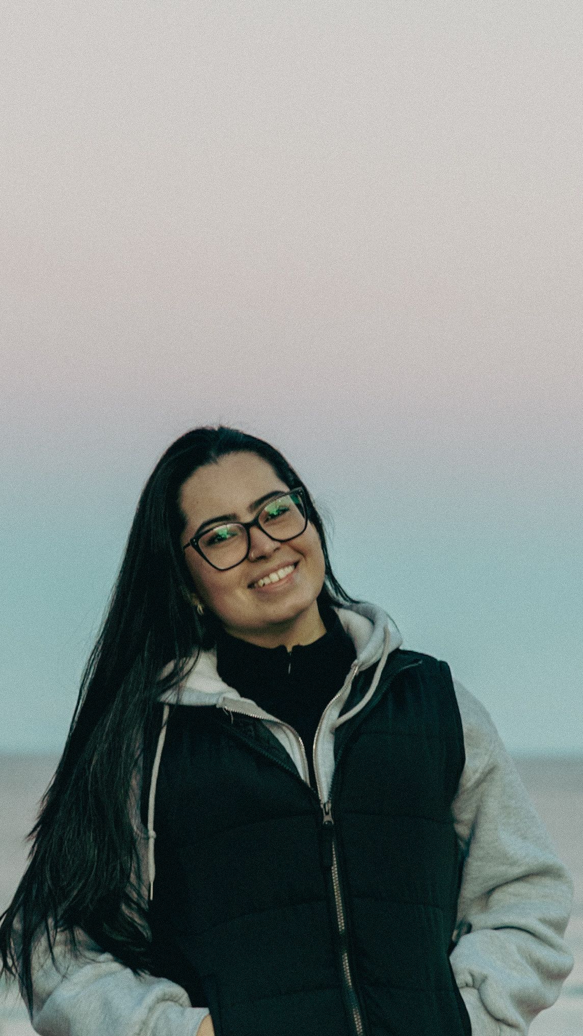 Smiling young woman with long black hair and glasses wearing a black puffer vest over a hoodie outdoors.