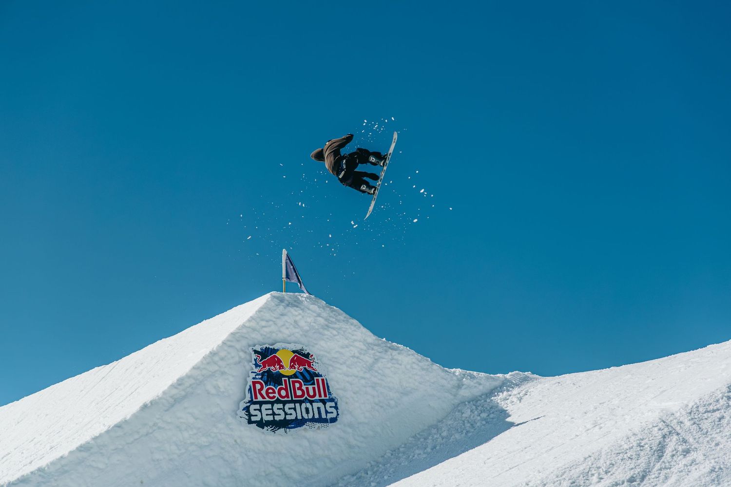 Snowboarder performs aerial trick over Red Bull Sessions branded snow jump against clear blue sky.