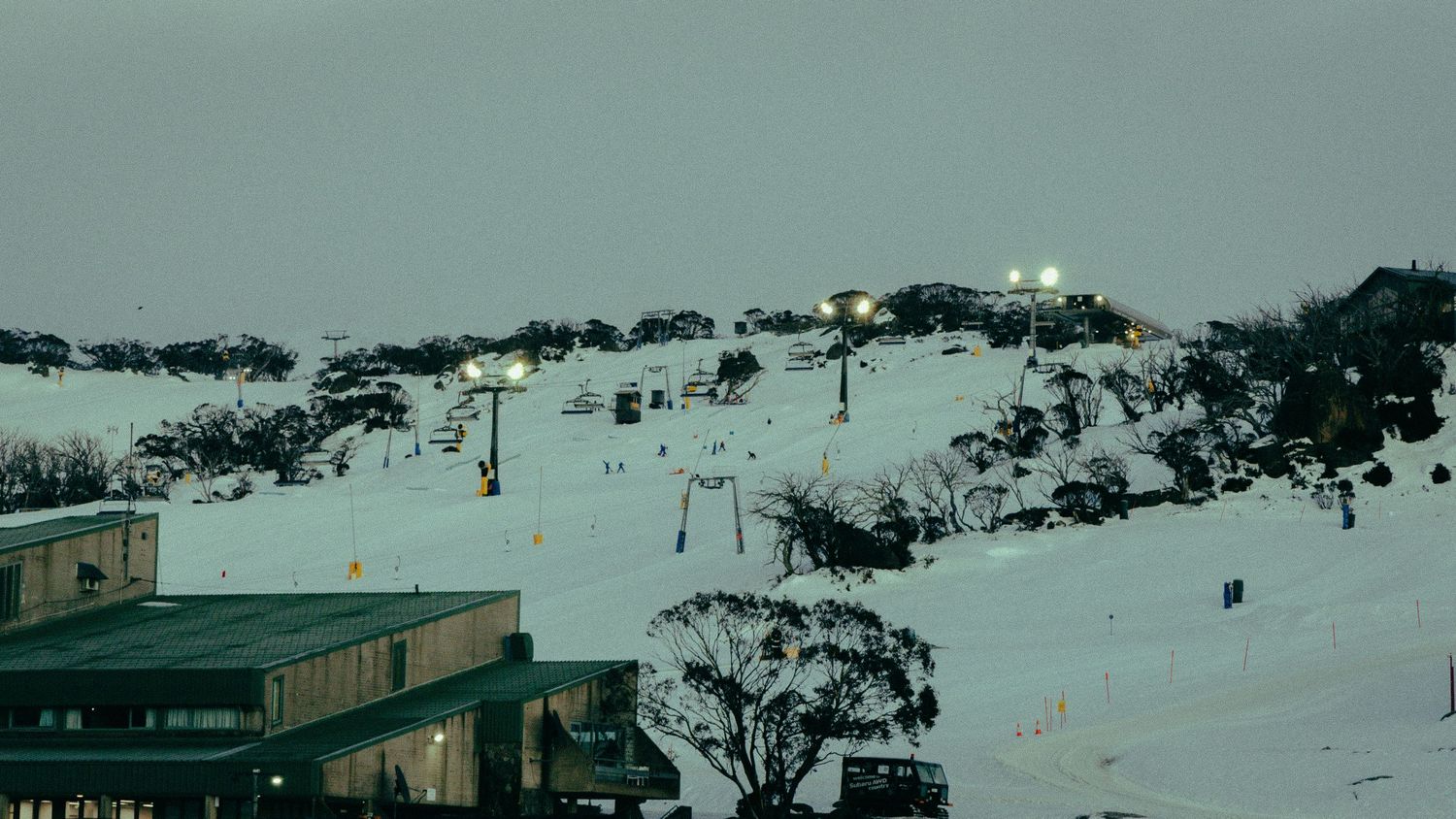 Snow-covered ski resort at dusk with illuminated slopes, ski lifts, and alpine lodge buildings in a winter mountain setting.
