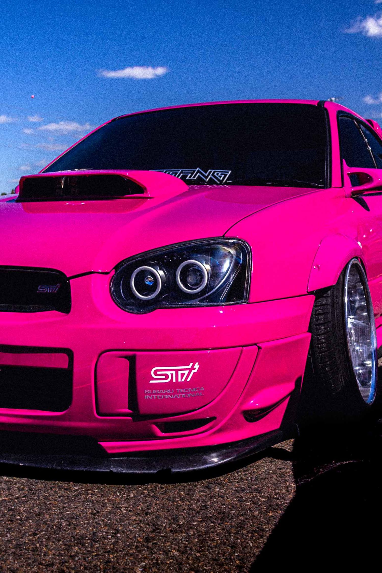Bright pink Subaru WRX STI with aftermarket headlights and lowered stance parked under a blue sky.