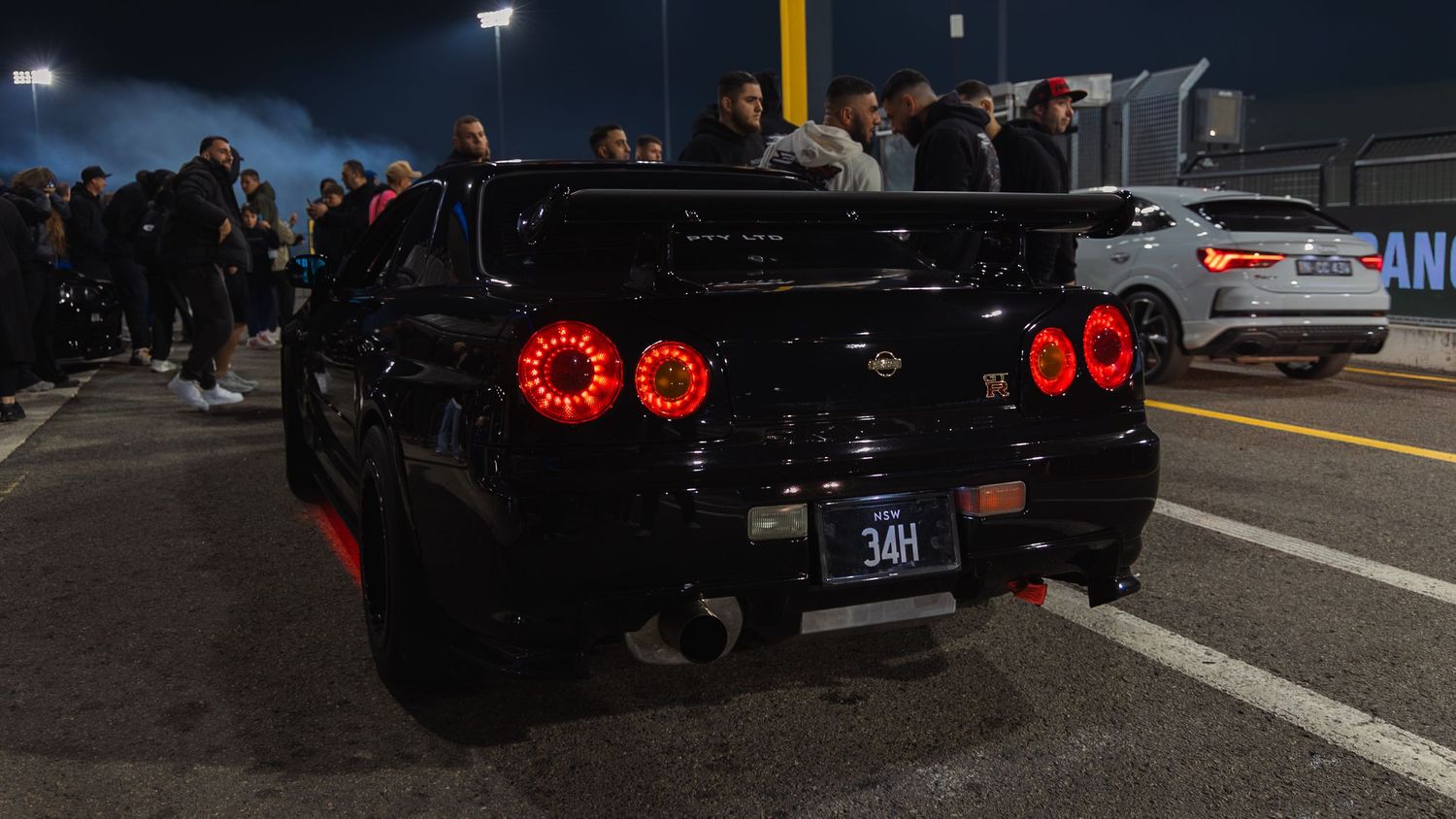 Black Nissan GT-R with red LED taillights at a nighttime car meet, license plate 34H visible.