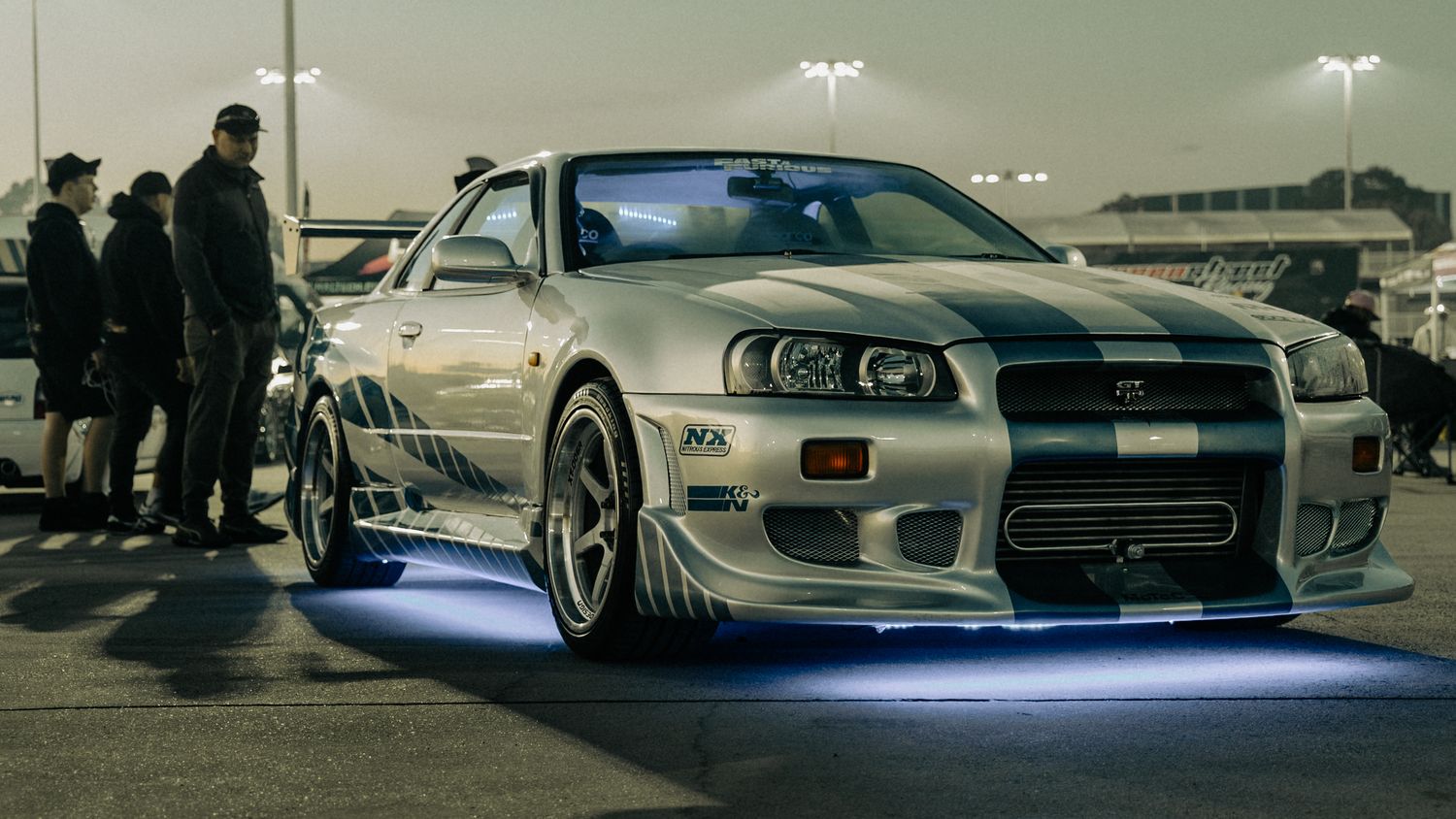A silver Nissan Skyline GT-R R34 with blue underglow lighting parked at a nighttime car meet.