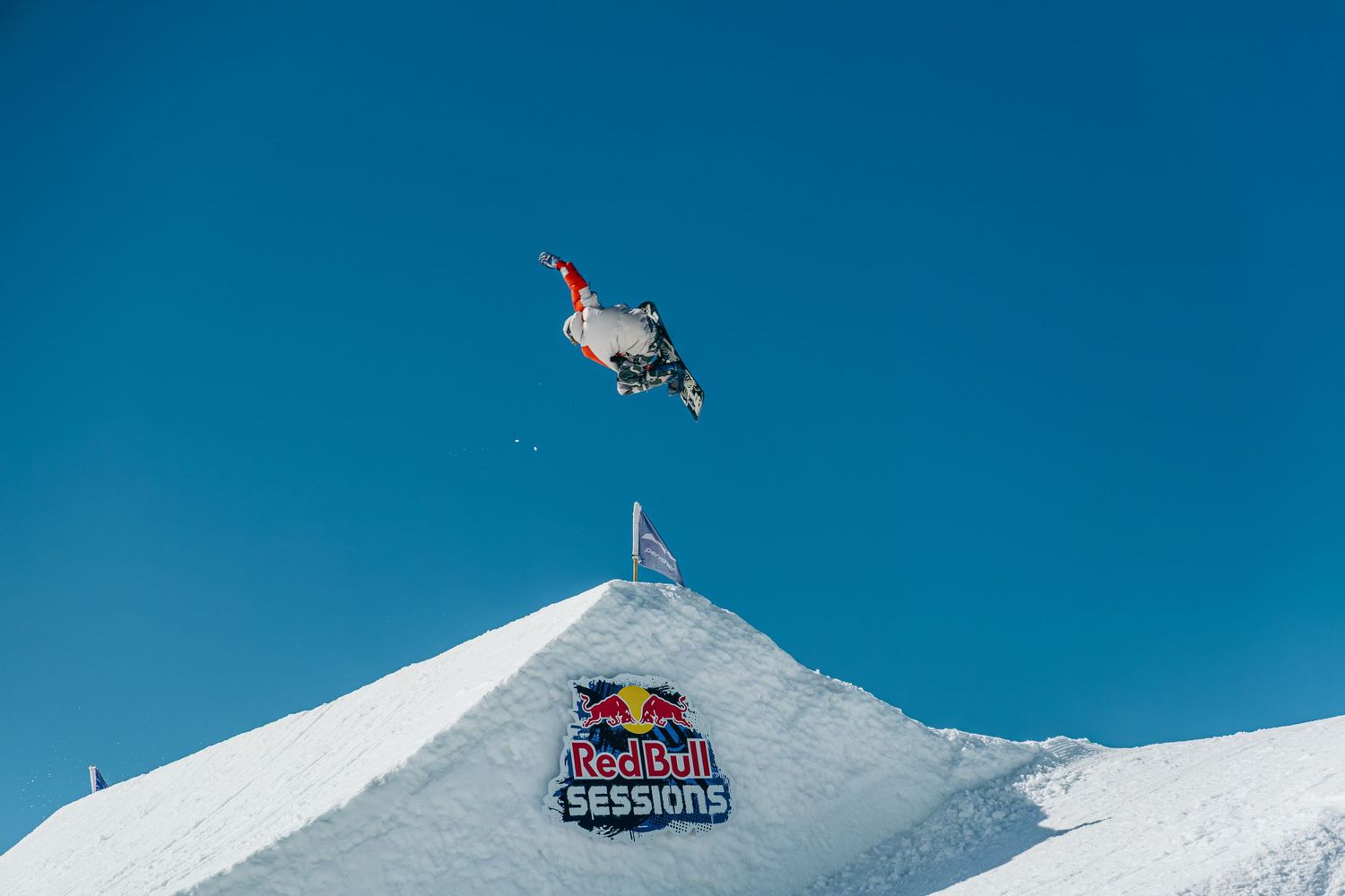 Snowboarder performs high aerial trick above Red Bull Sessions branded snow ramp against clear blue sky.