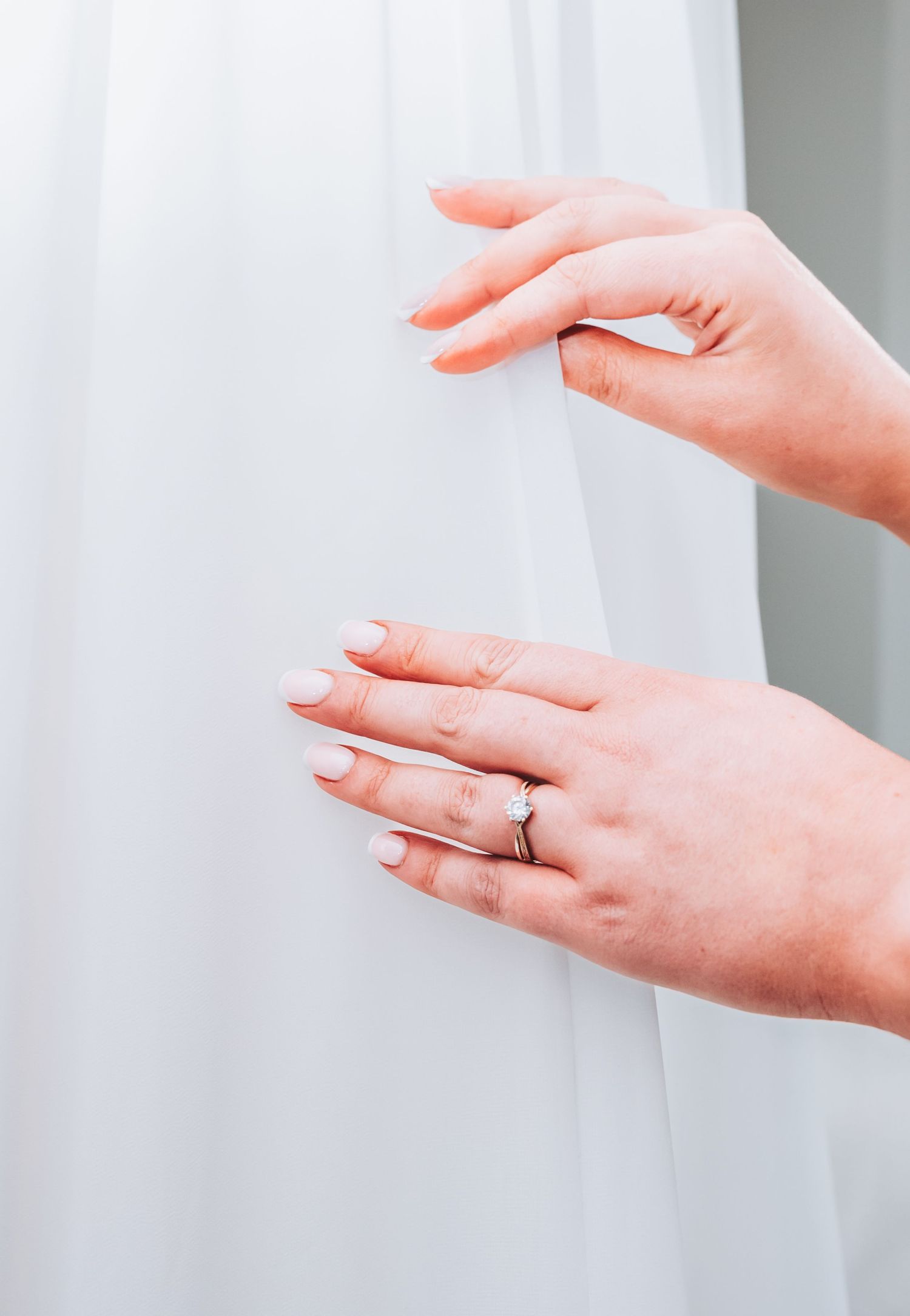 Close-up of bride's hands with white manicure and wedding ring delicately holding a flowing white bridal gown.