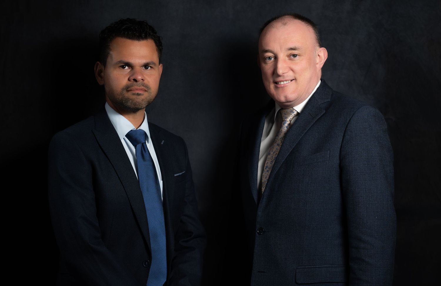 Two professional men in dark suits and ties pose for a formal business portrait against a dark background.