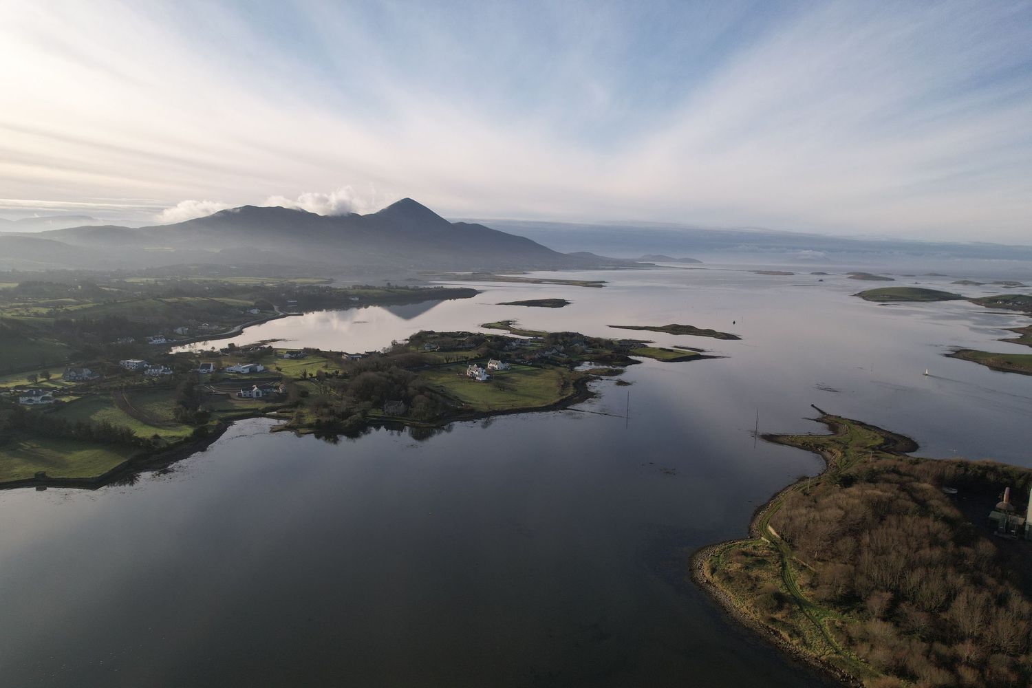 Aerial view of a serene Irish lake with scattered islands and a mountain backdrop under soft clouds.