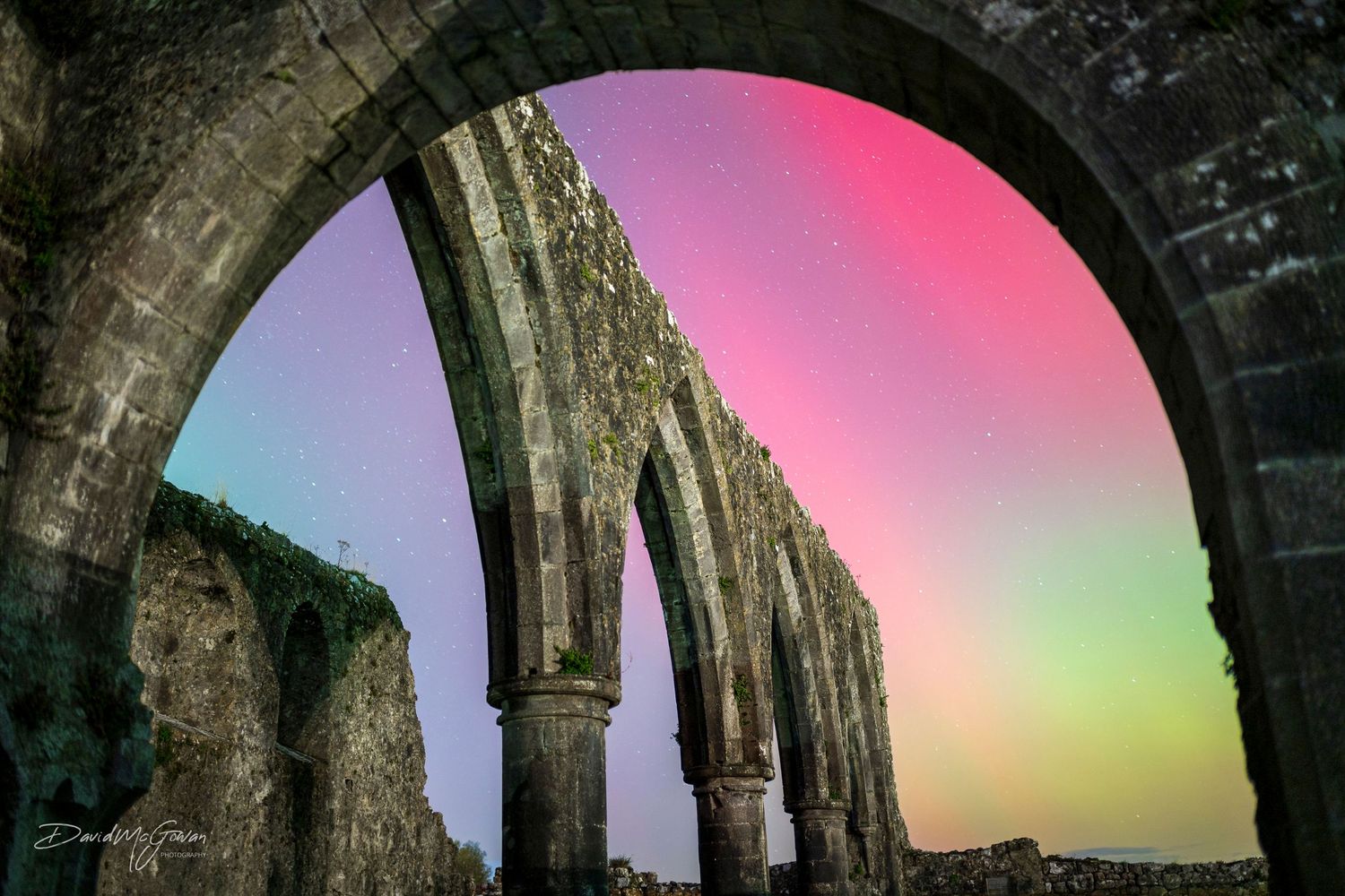 Colorful aurora borealis glowing pink and green through the arches of an ancient stone ruin.