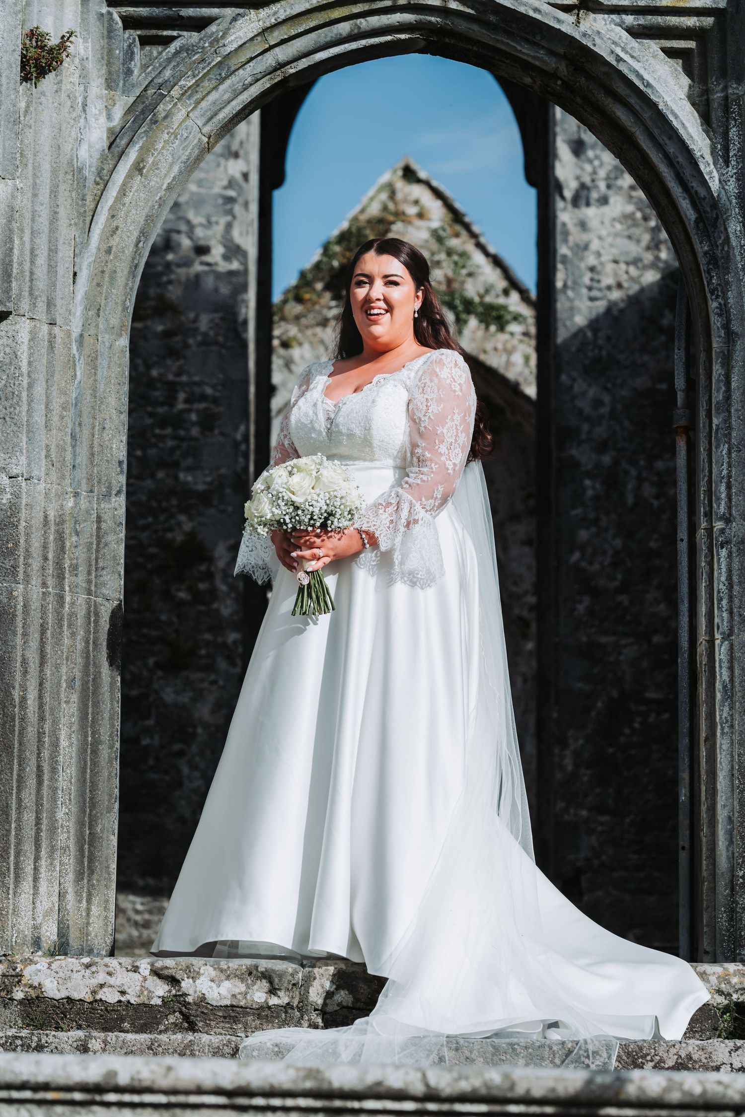 Bride in elegant long-sleeve lace wedding dress holding bouquet, posing in ancient stone archway.