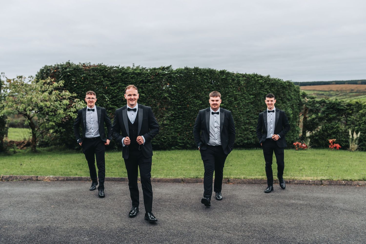Four groomsmen in black suits standing socially distanced on a driveway.