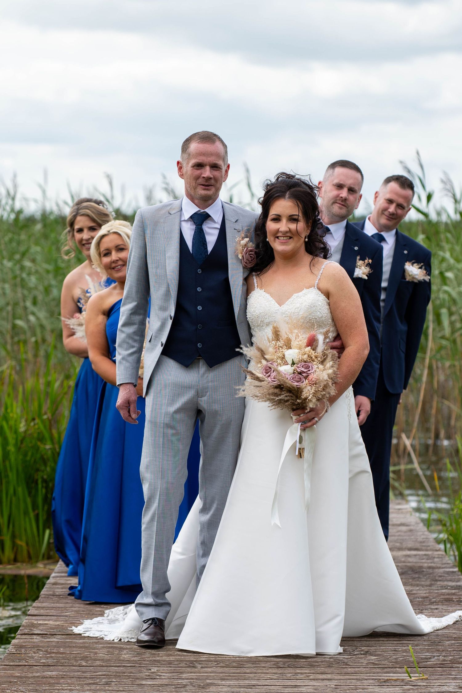Wedding party poses on a wooden dock, bride in white gown holding bouquet with groom in gray suit and guests behind.