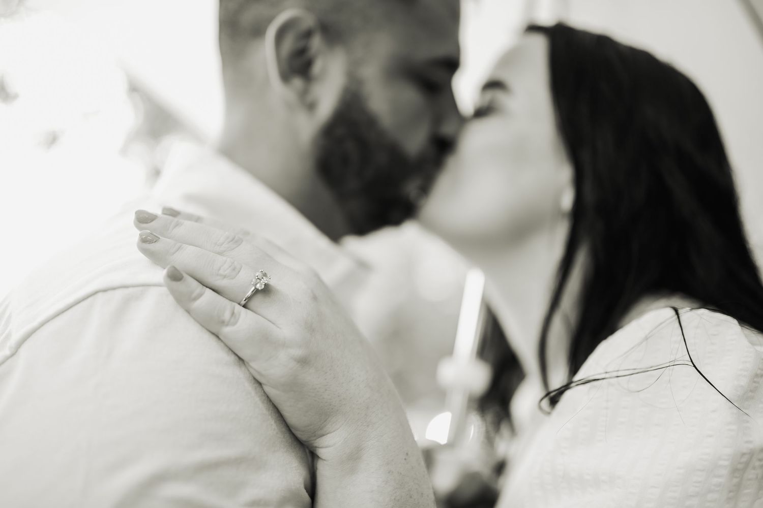 Black and white photo of a couple sharing a romantic kiss, woman displaying an engagement ring on her hand.