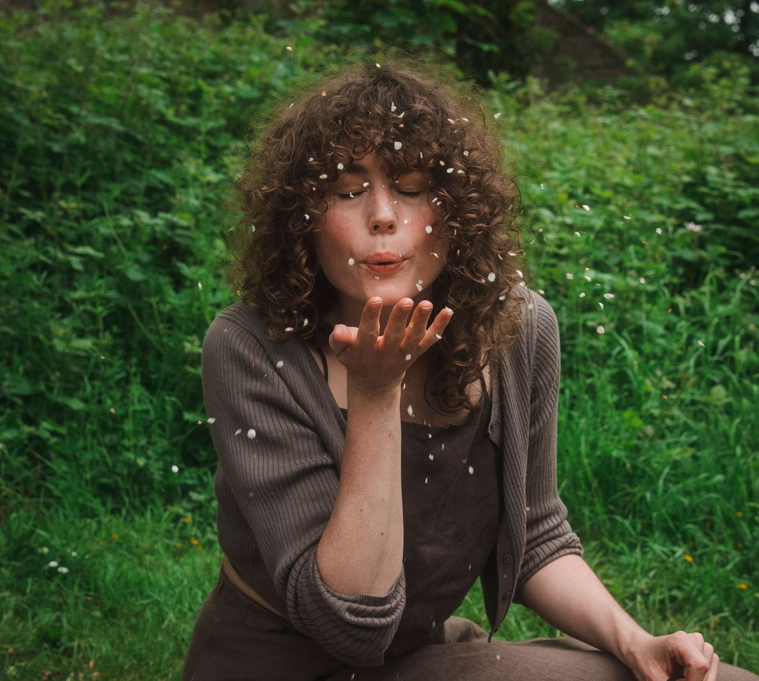 Curly-haired woman blowing confetti petals from her hands while sitting outdoors in a lush green garden.