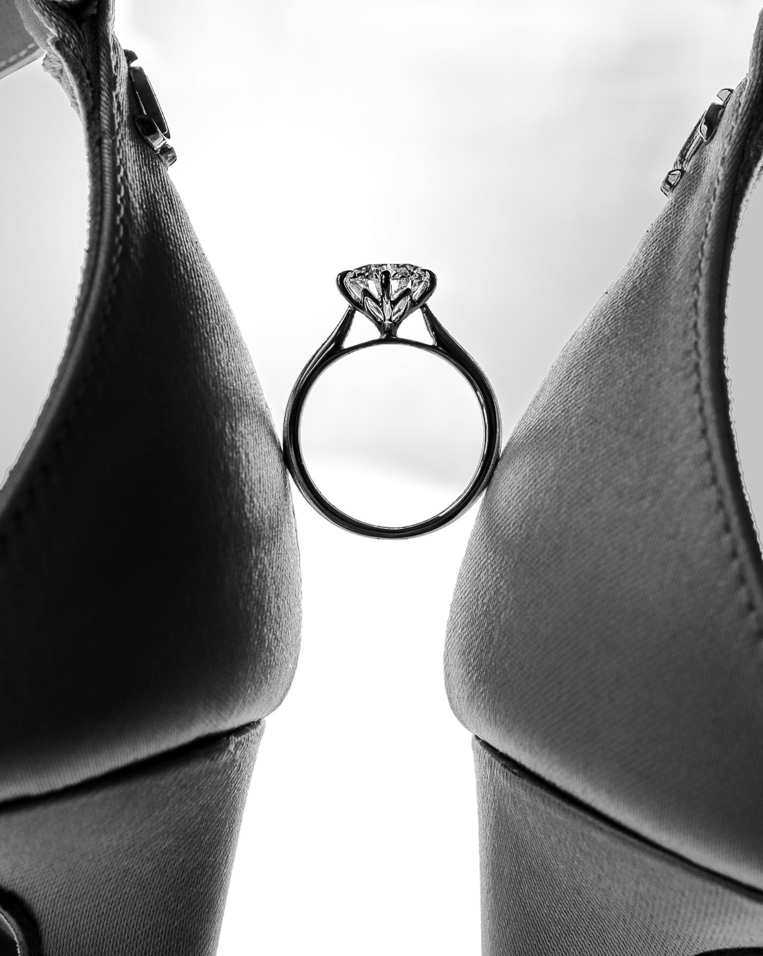 Black and white photo of a diamond solitaire engagement ring balanced between two high heel shoes.