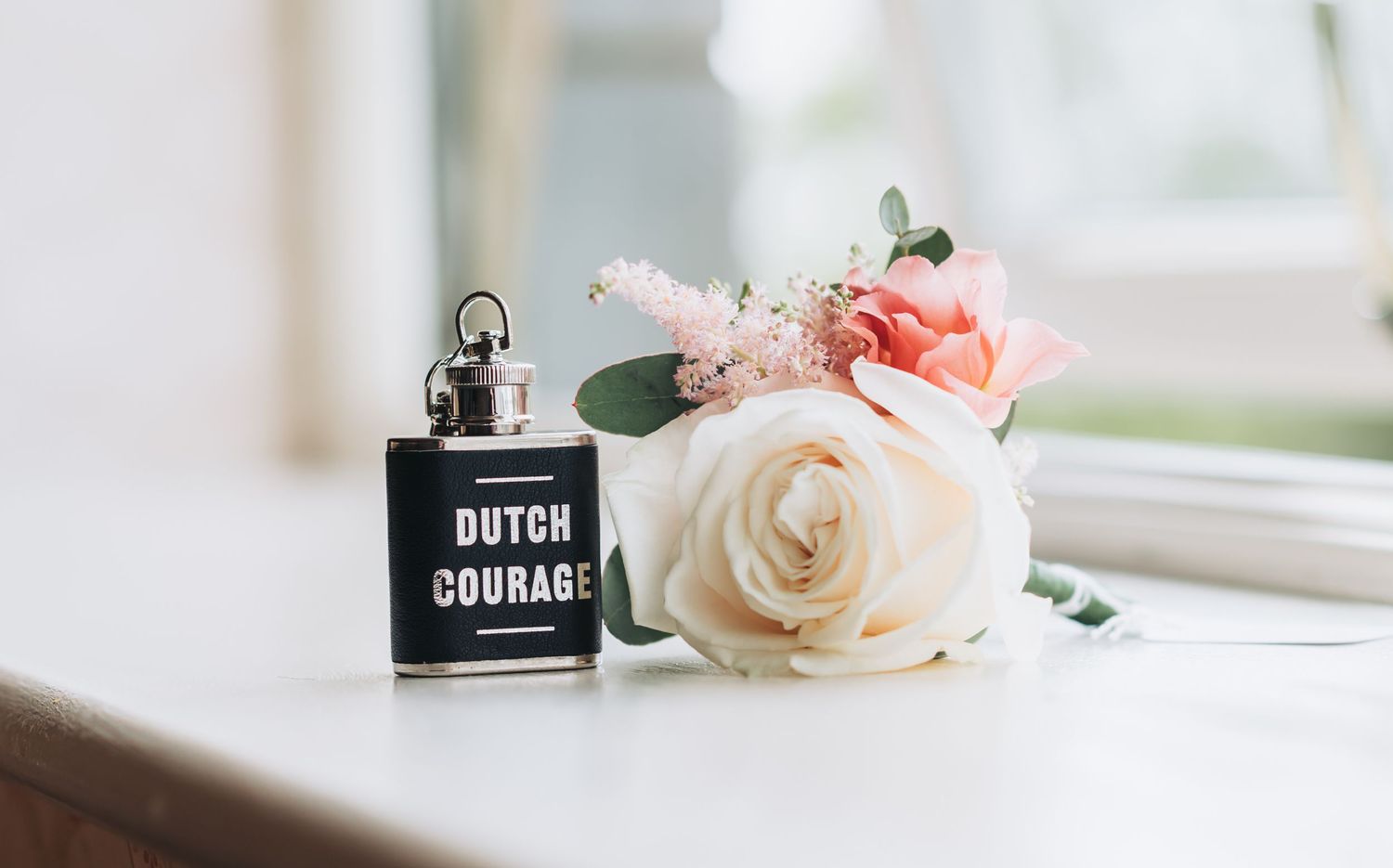 A black Dutch Courage flask beside a white and pink rose boutonniere on a white windowsill.