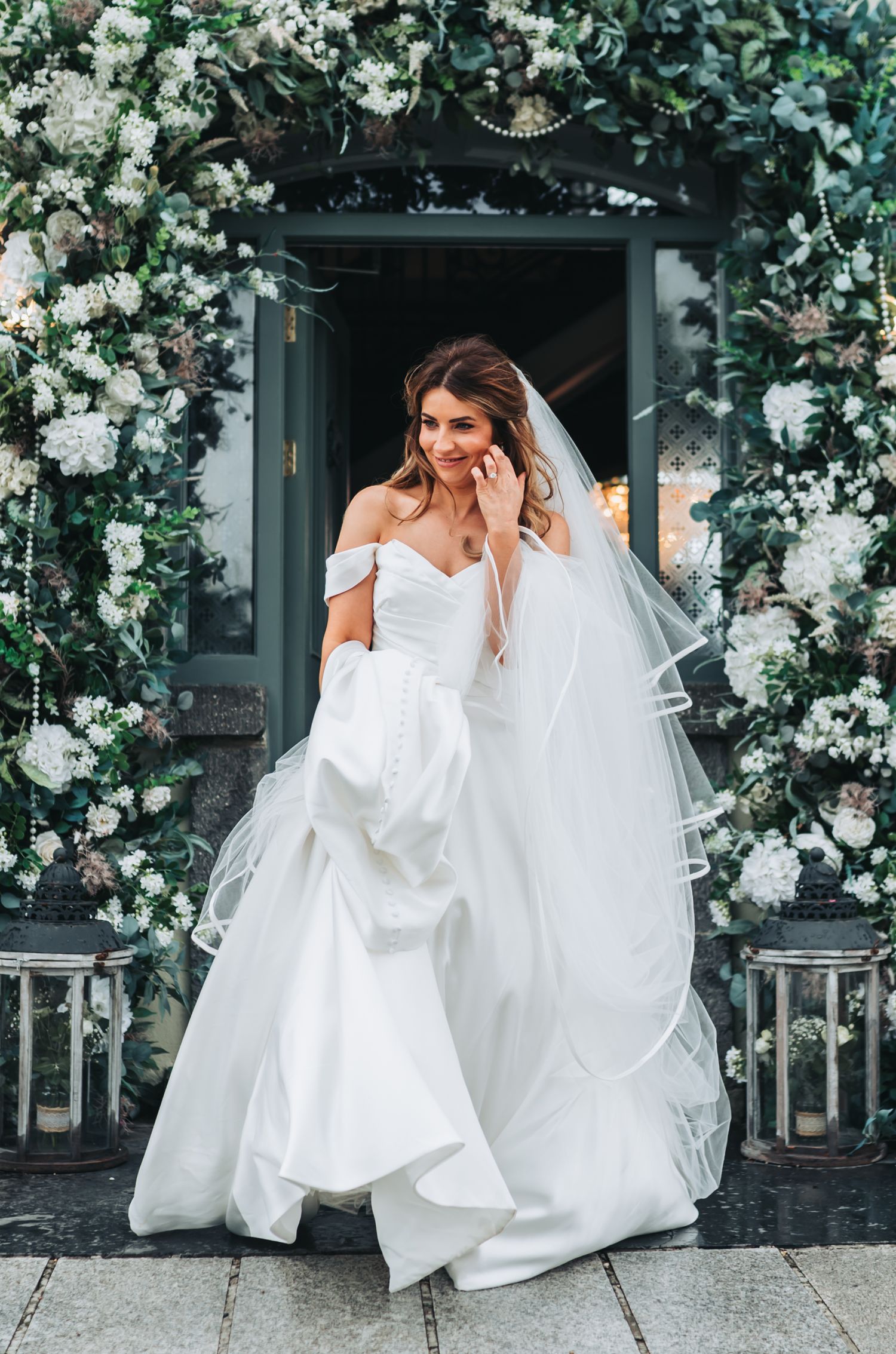 Bride in off-shoulder white gown with veil standing in flower-adorned doorway on wedding day.