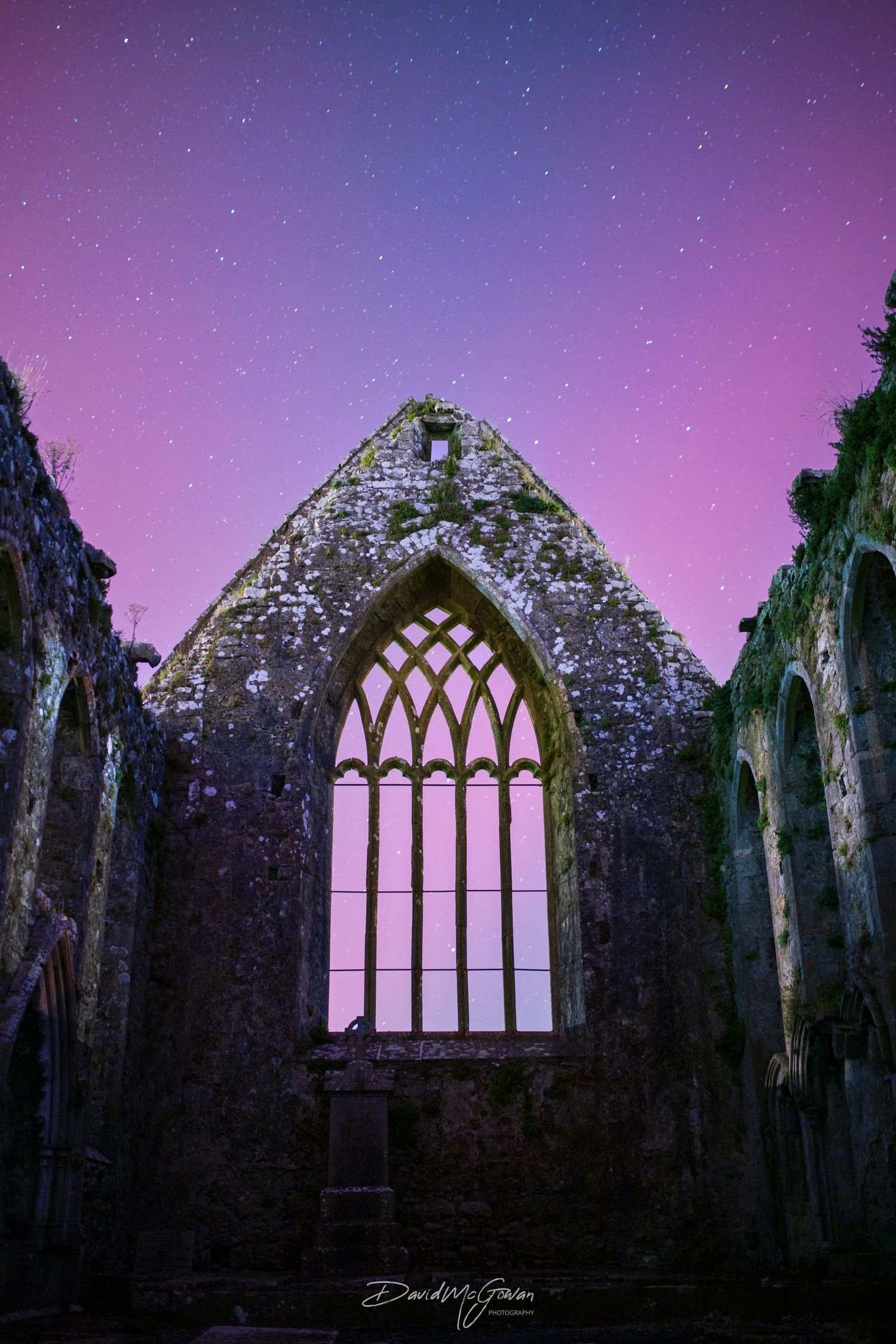 Ancient stone church ruins with Gothic arched window under a stunning purple starry night sky.