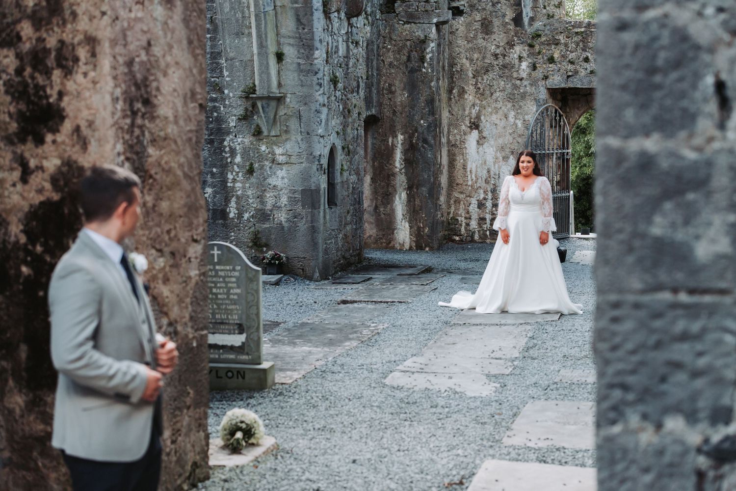 Bride in white gown approaches groom among ancient stone ruins during romantic wedding first look.