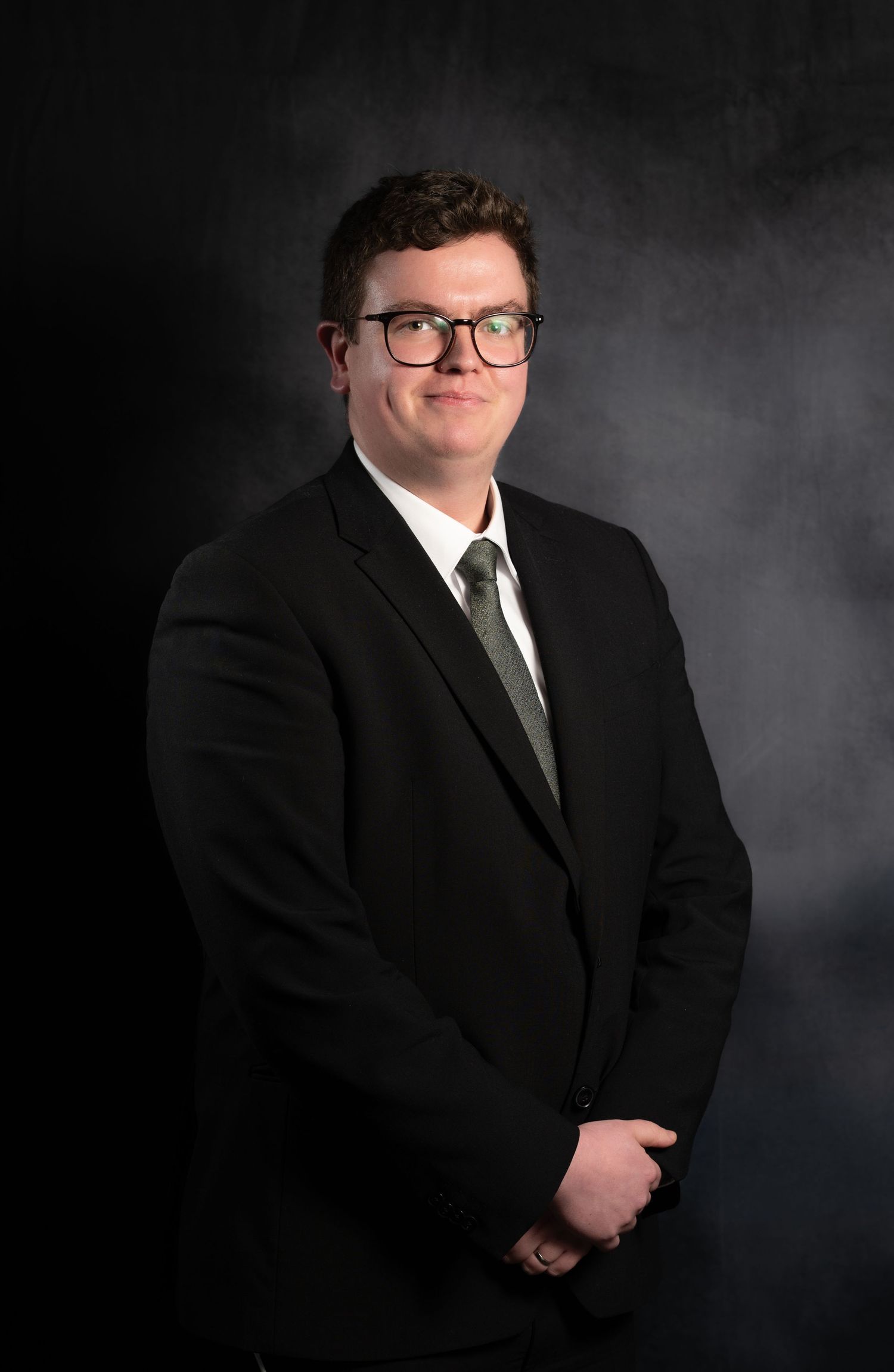 Young man in black suit and green tie with glasses smiling for professional headshot photo.