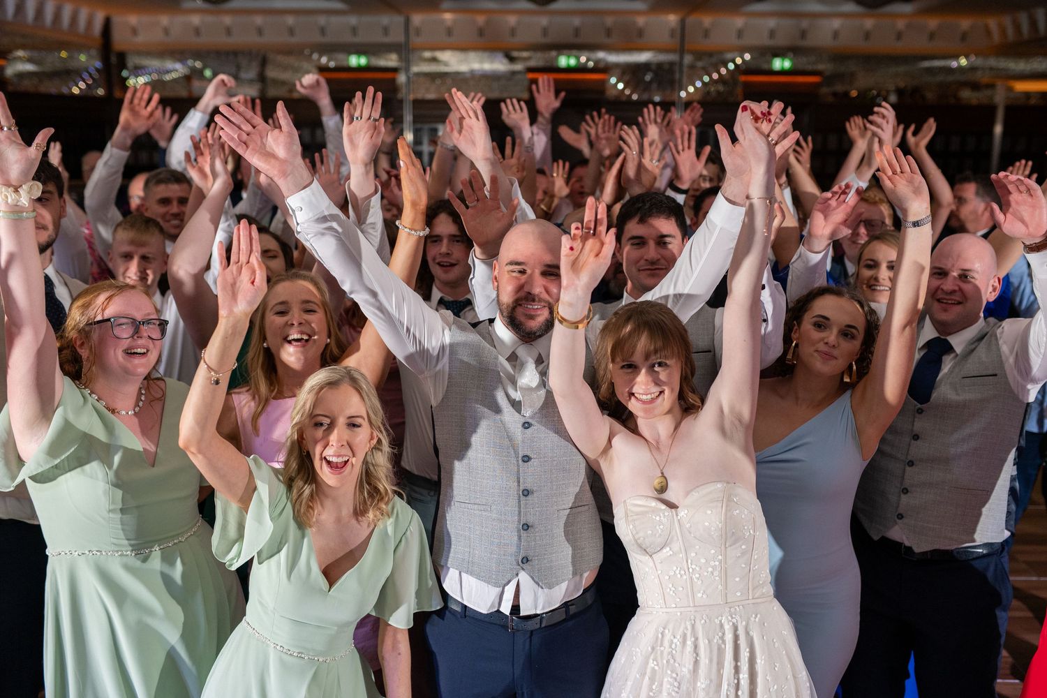 Joyful wedding celebration with bride, groom, and guests raising hands together on dance floor.