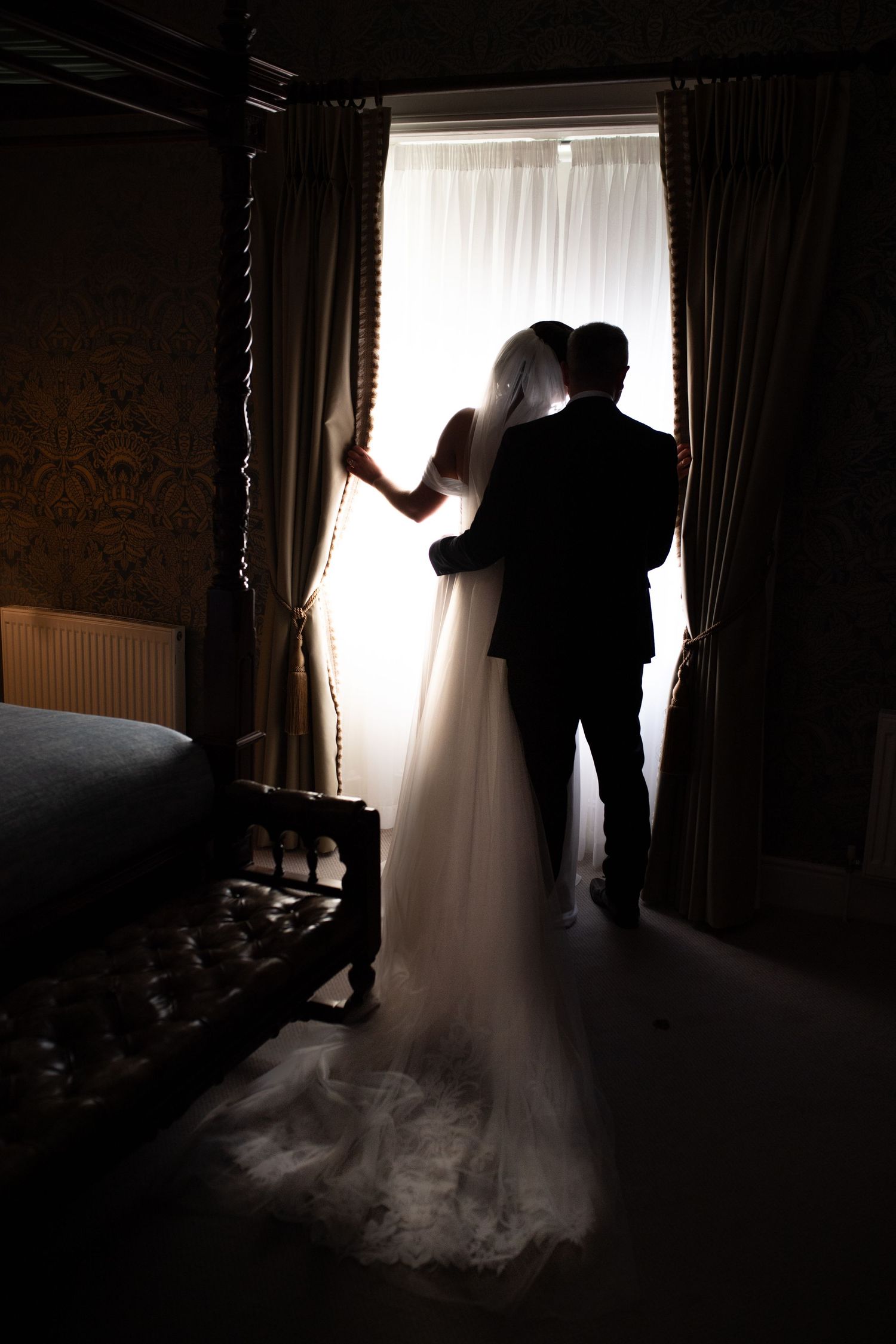 Silhouetted couple standing by window in elegant room with flowing wedding dress train.