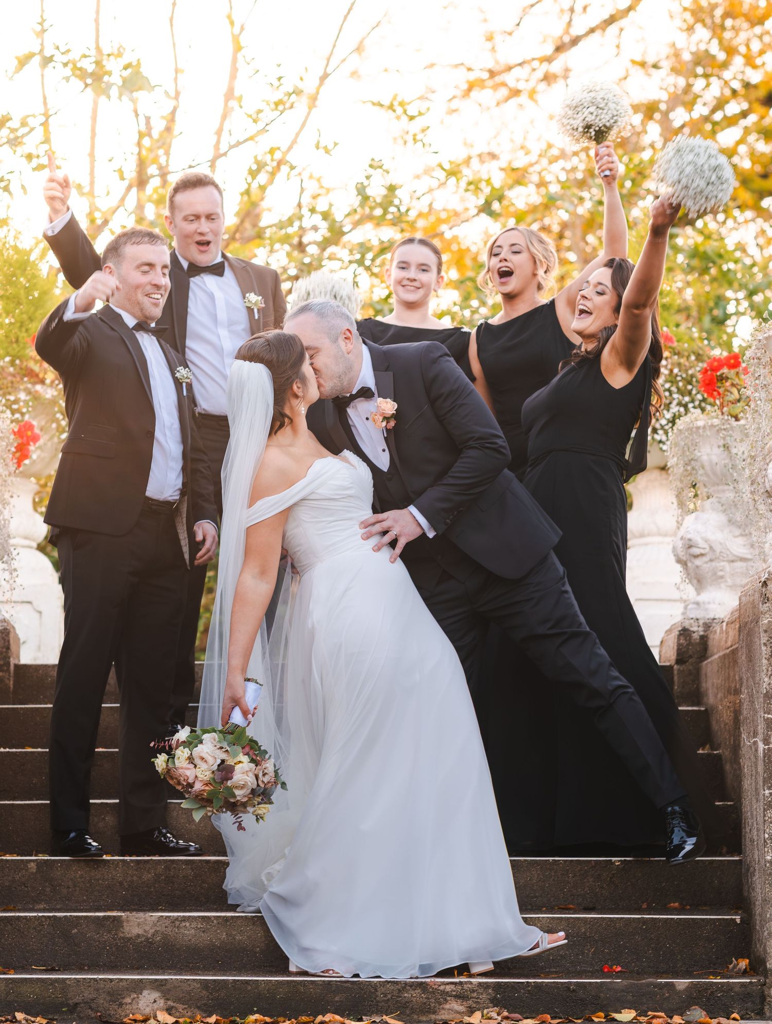 Wedding party celebrates on stone steps as bride and groom share a romantic dip kiss in autumn setting.