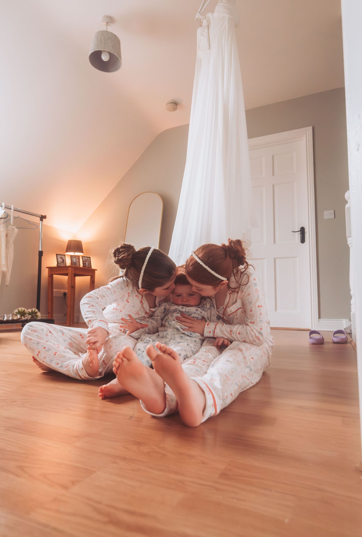 Two young sisters in matching pajamas sit on hardwood floor sharing a tender moment with baby sibling.