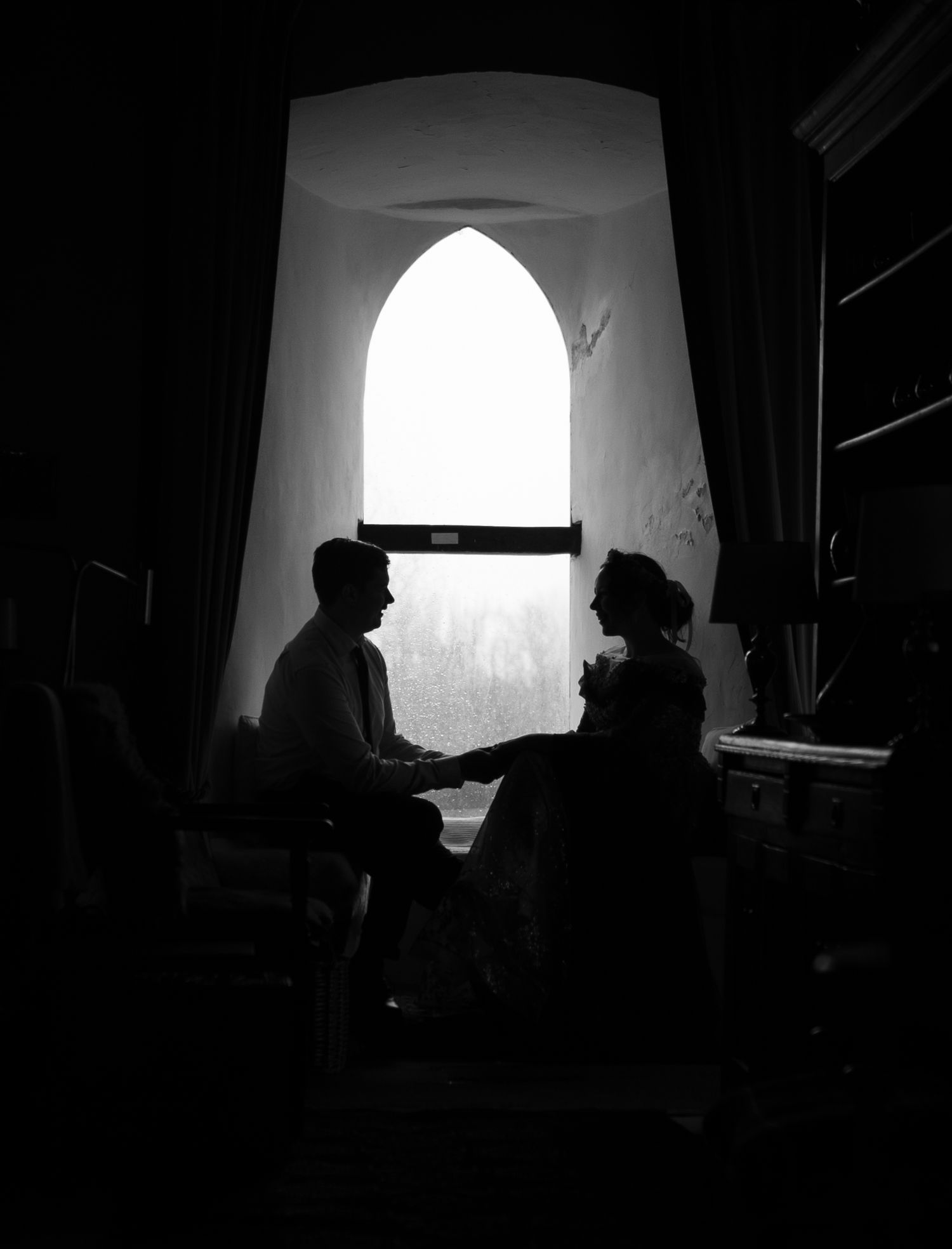 Black and white silhouette of two people conversing by a Gothic arch window in a dimly lit room.