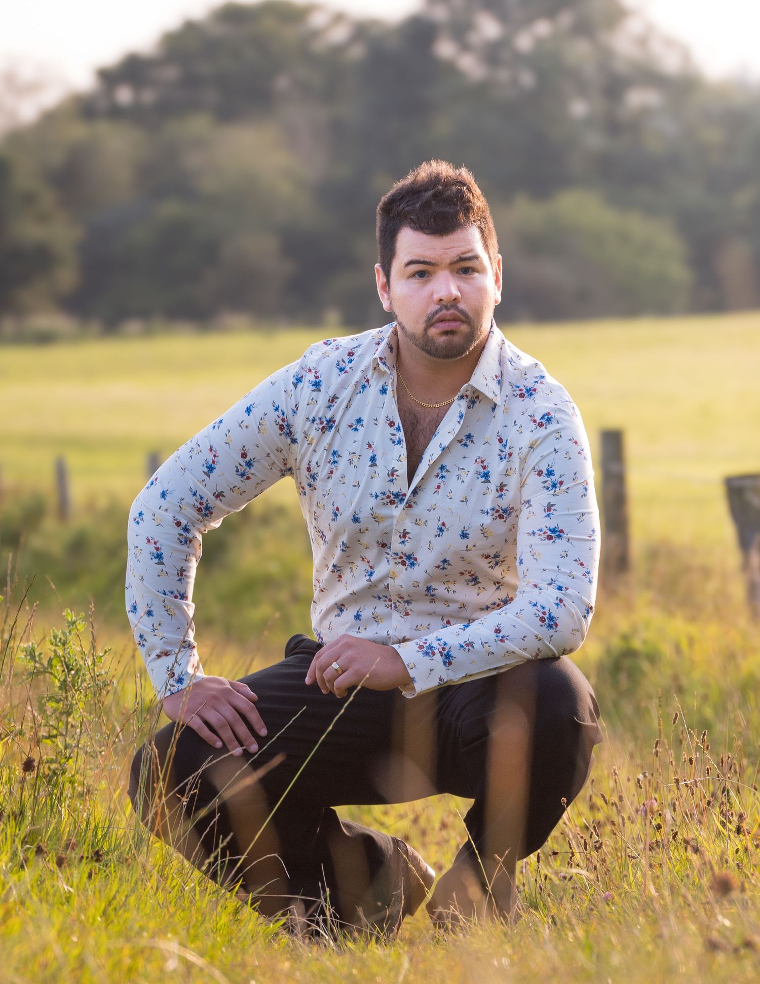 Man in floral shirt crouching in green field during golden hour outdoors.