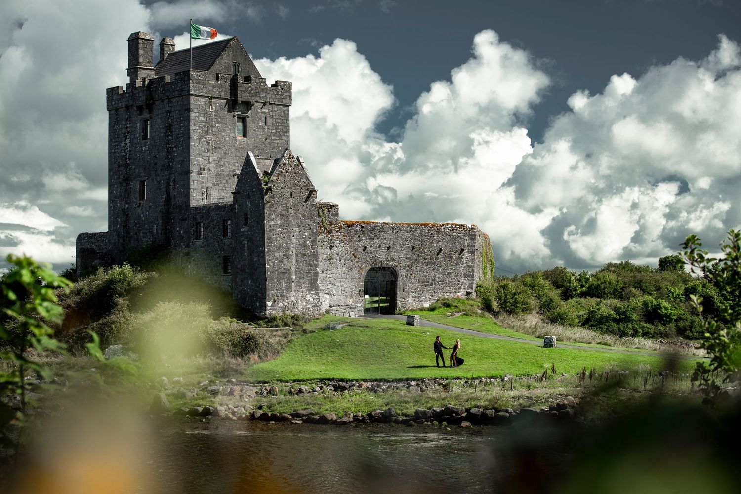 Ancient Irish stone castle with green lawn, flying flag, surrounded by water under dramatic cloudy sky.
