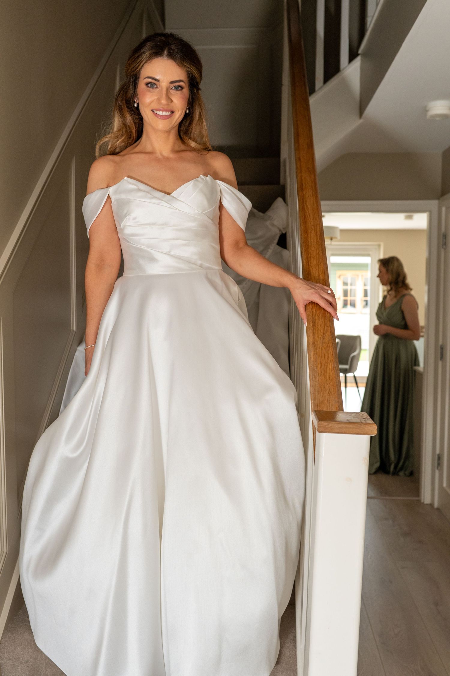 Smiling bride in off-shoulder satin wedding gown poses on staircase with bridesmaid in background.