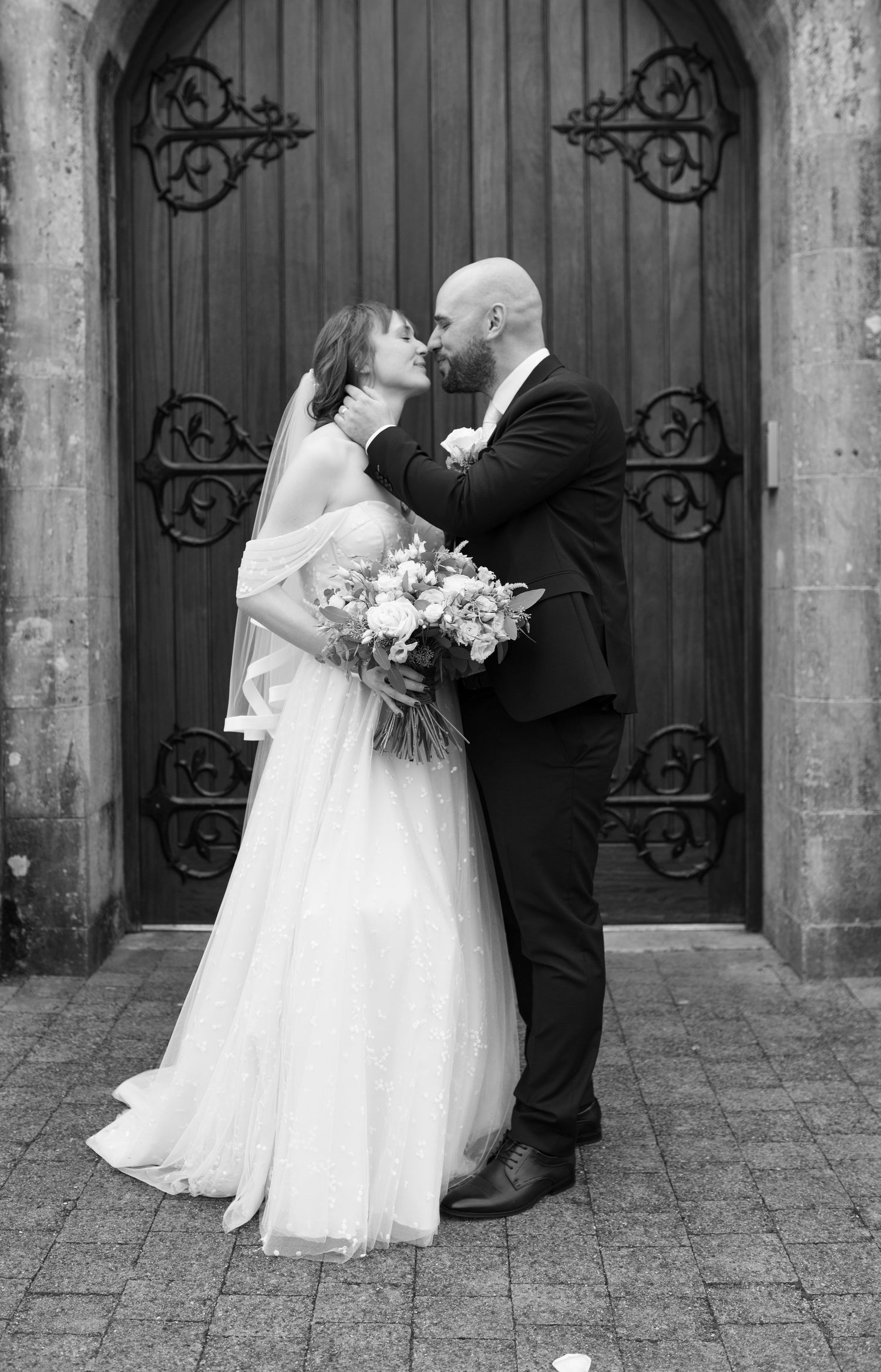 Bride and groom share a kiss outside ornate wooden church doors, bride holding a floral bouquet.