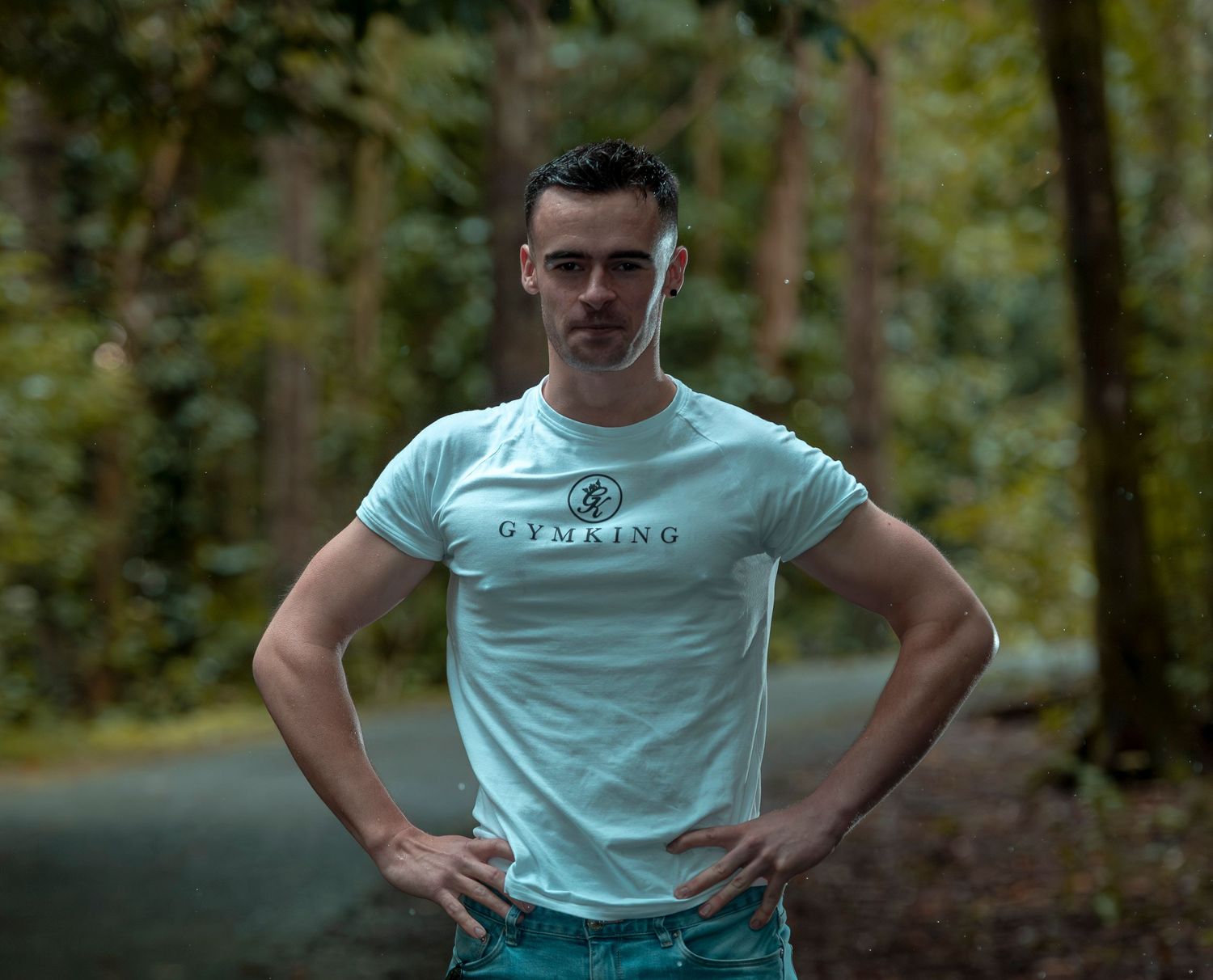 Muscular man in mint green Gymking t-shirt posing with hands on hips on a forest road.