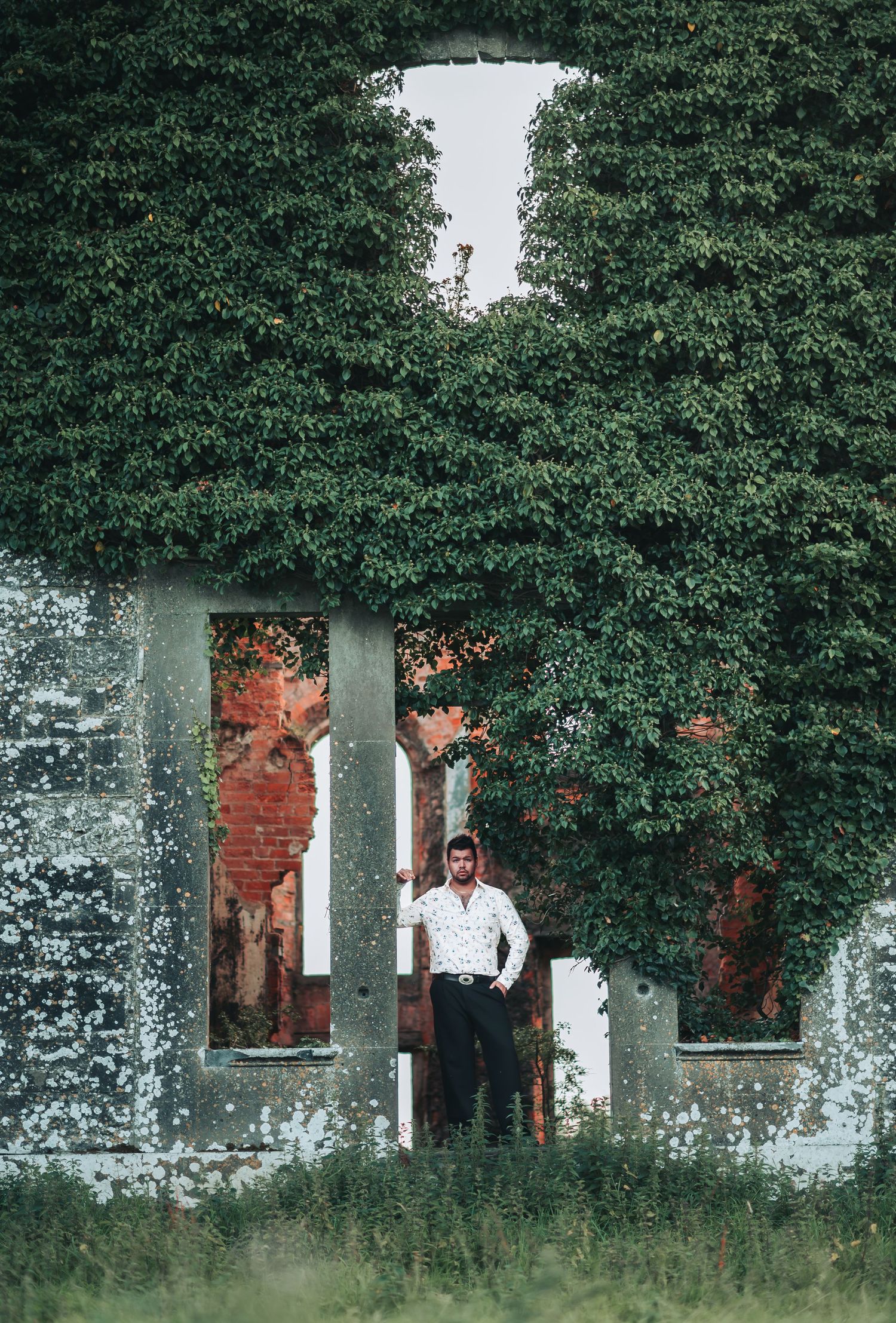 Man standing in ivy-covered ruins of an old brick building with overgrown walls and open window frames.