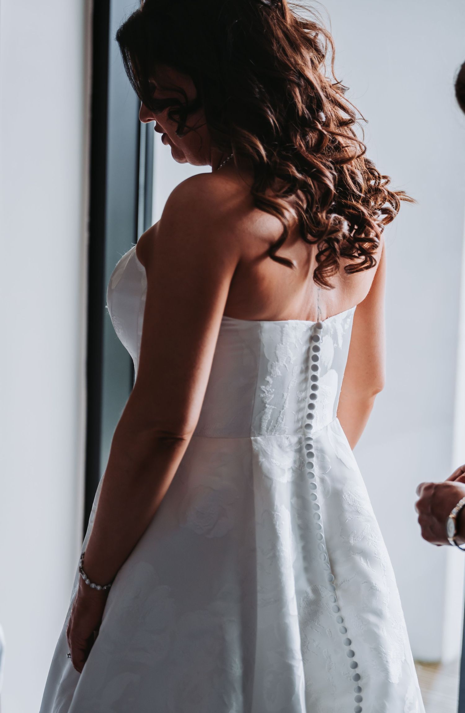 Bride in a white strapless wedding gown with button back detail, curly brown hair, being helped to dress.