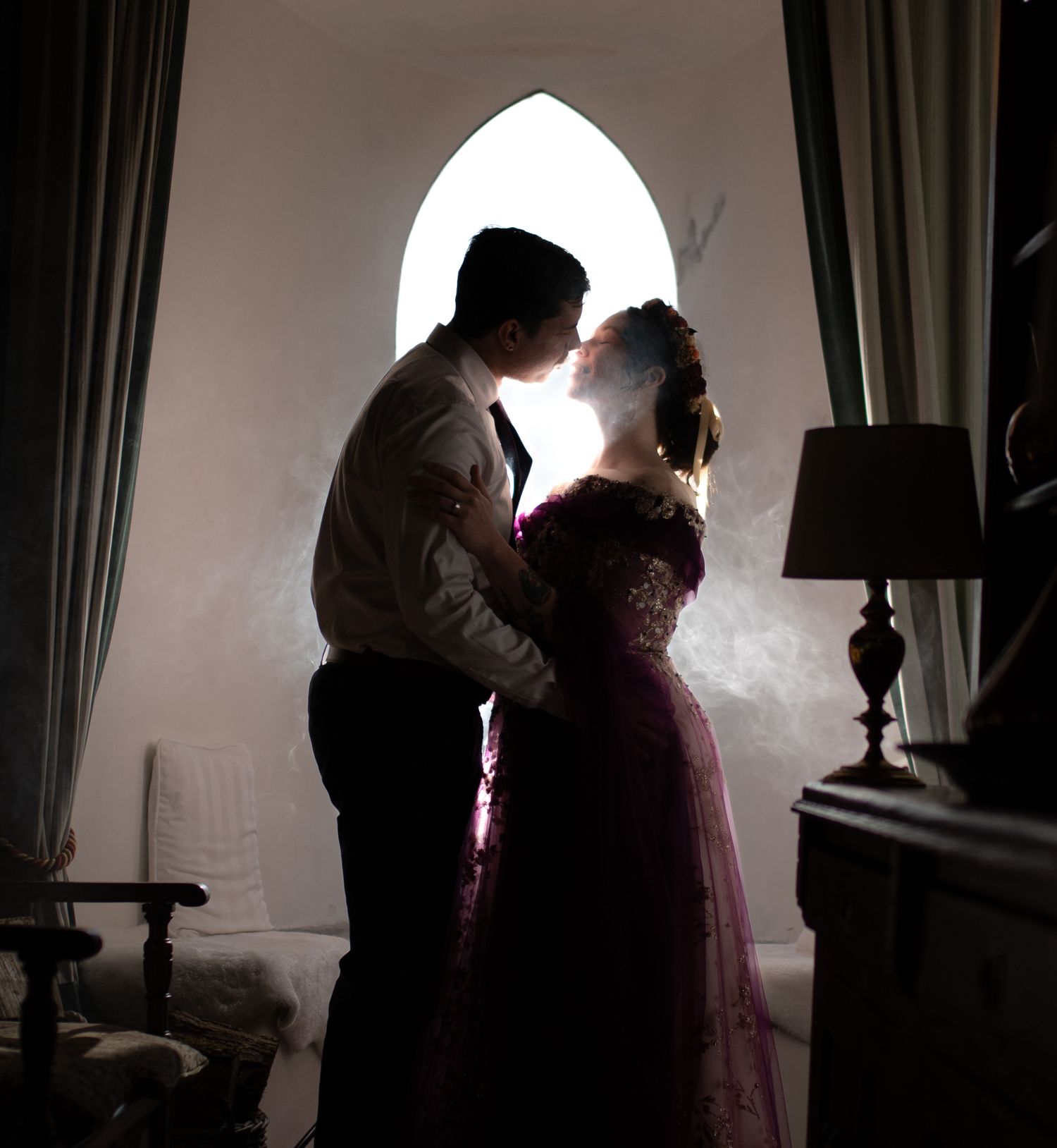 Couple share romantic kiss silhouetted against gothic arched window in dimly lit castle interior.