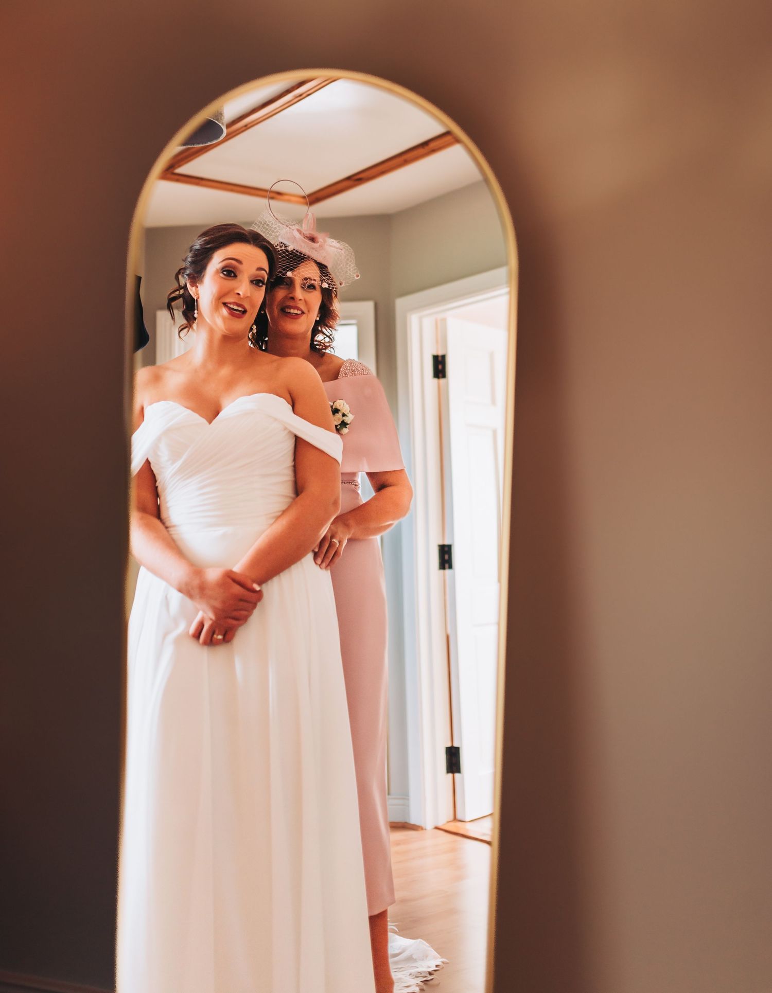 Bride in white off-shoulder gown reflected in arched mirror with bridesmaid in pink suit helping her prepare.